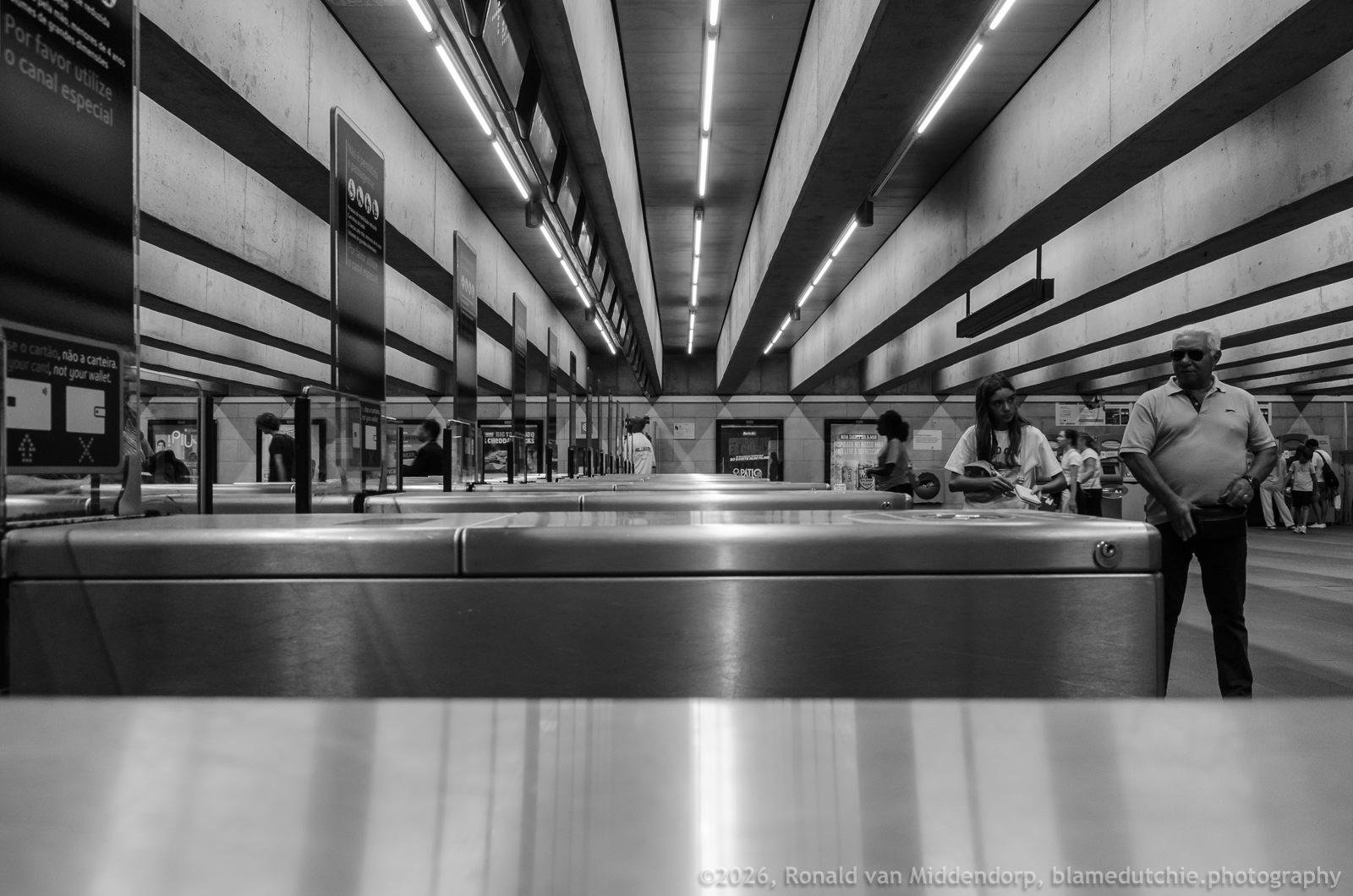 Low-angle black-and-white view of a subway station hall with metal turnstiles in the foreground, long ceiling light strips, and commuters standing and walking near the gates, including a man in sunglasses on the right.