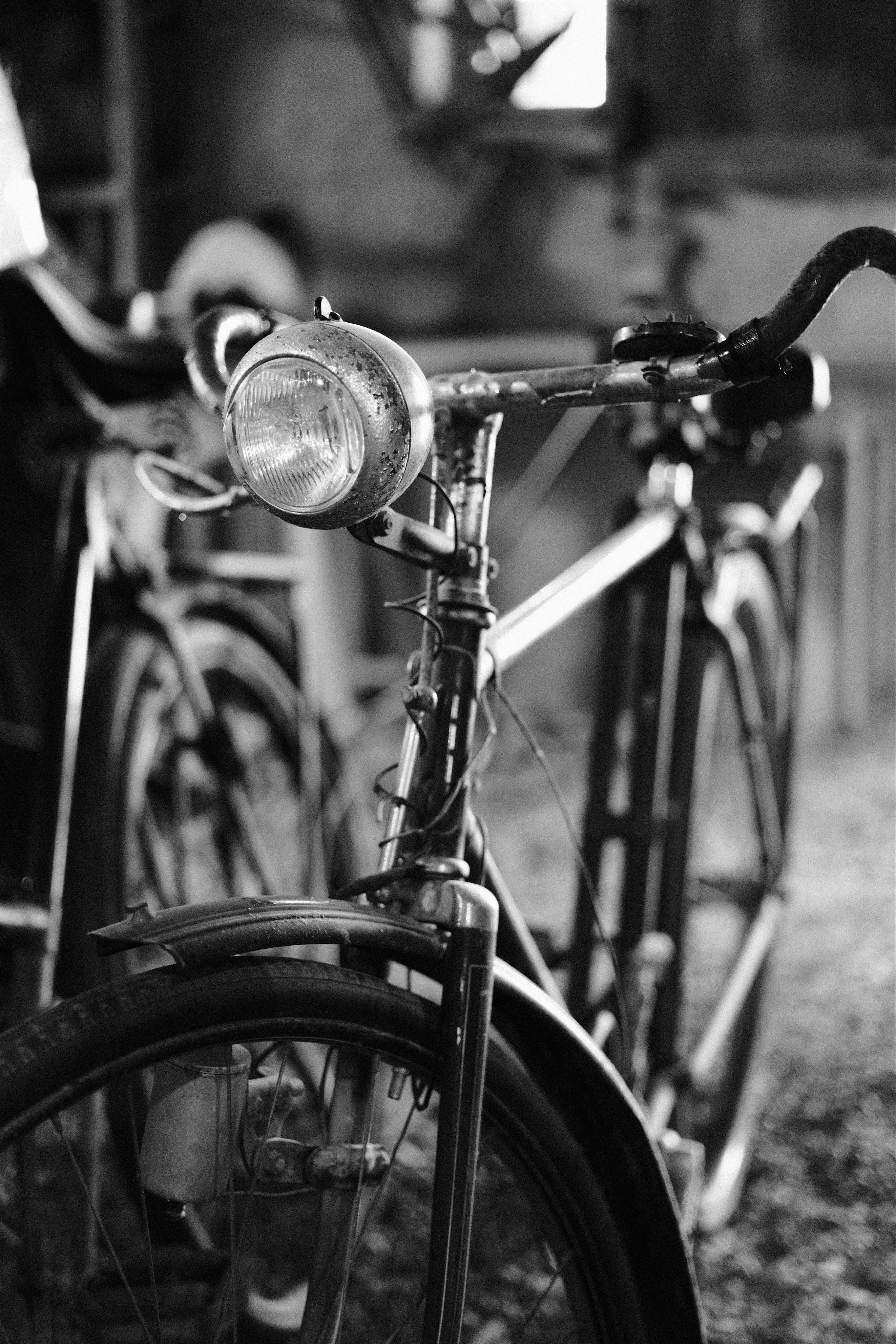 Close-up black-and-white photo of a vintage bicycle’s front wheel, fender, and handlebar-mounted headlight in the foreground.