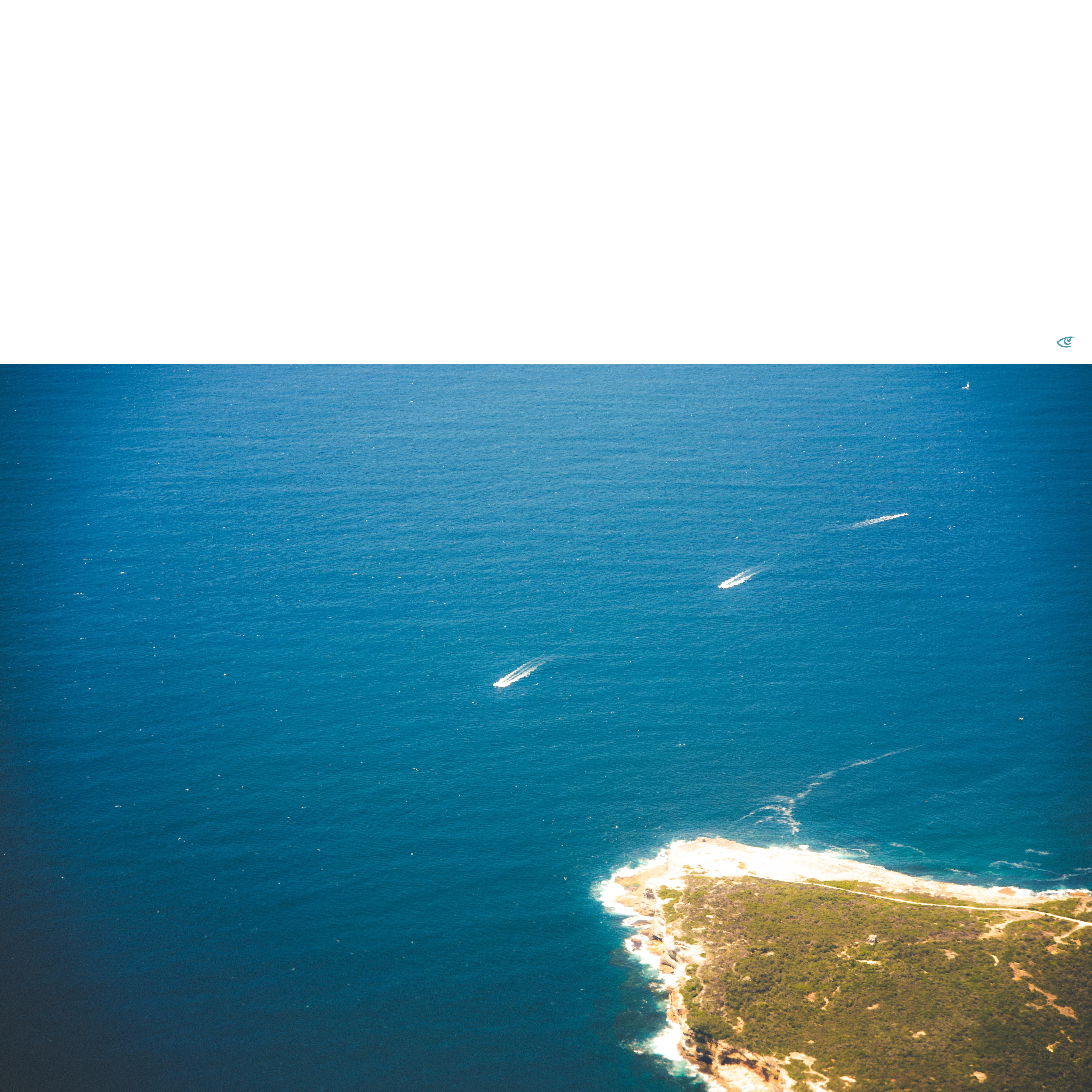 Aerial view of deep blue ocean with several small white boats leaving wakes, and a rocky green peninsula with a pale sandy shoreline in the lower right corner.