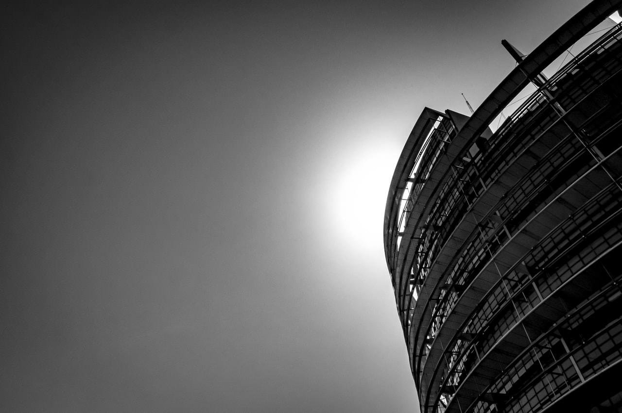 Curved modern building with multiple floors and rows of windows, viewed from a low angle. Bright sunlight creates a halo effect behind the structure, emphasizing its sleek architectural lines against the clear sky. Black and white composition enhances contrast and details.