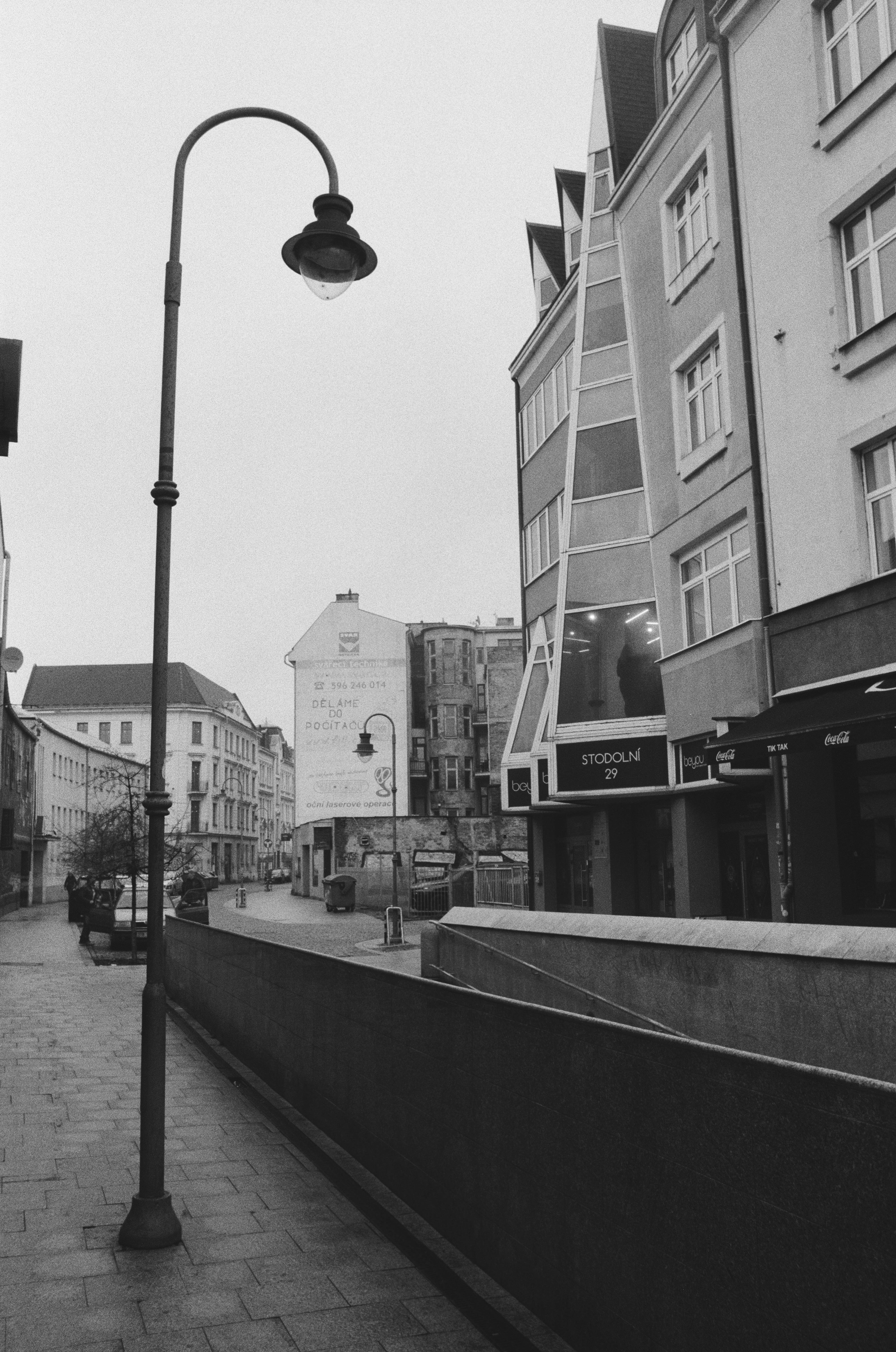 The image is a black and white photo of a street scene. It shows a sidewalk with a street lamp in the foreground and buildings lining the street. There is a prominent modern building with large windows on the right side, displaying the address "Stodolni 29." In the background, there is a billboard on a building wall. The atmosphere seems quiet and possibly early in the day or during an overcast weather condition.