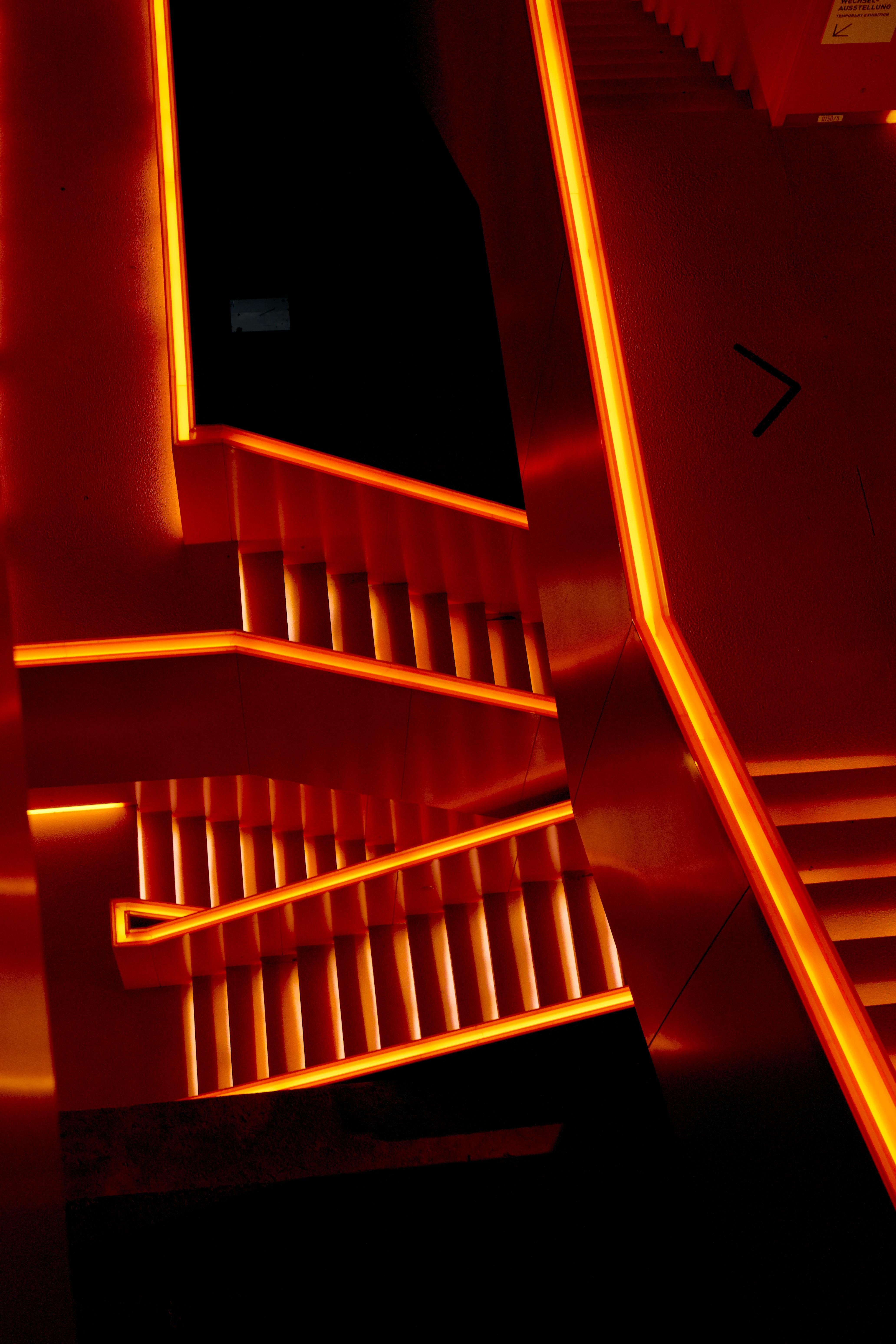 Stairs with bright orange-red neon lights lining the edges, creating a zigzag pattern as they ascend. The surrounding walls and steps are illuminated by the glow, contrasting with the dark areas. An arrow sign is visible on the wall.