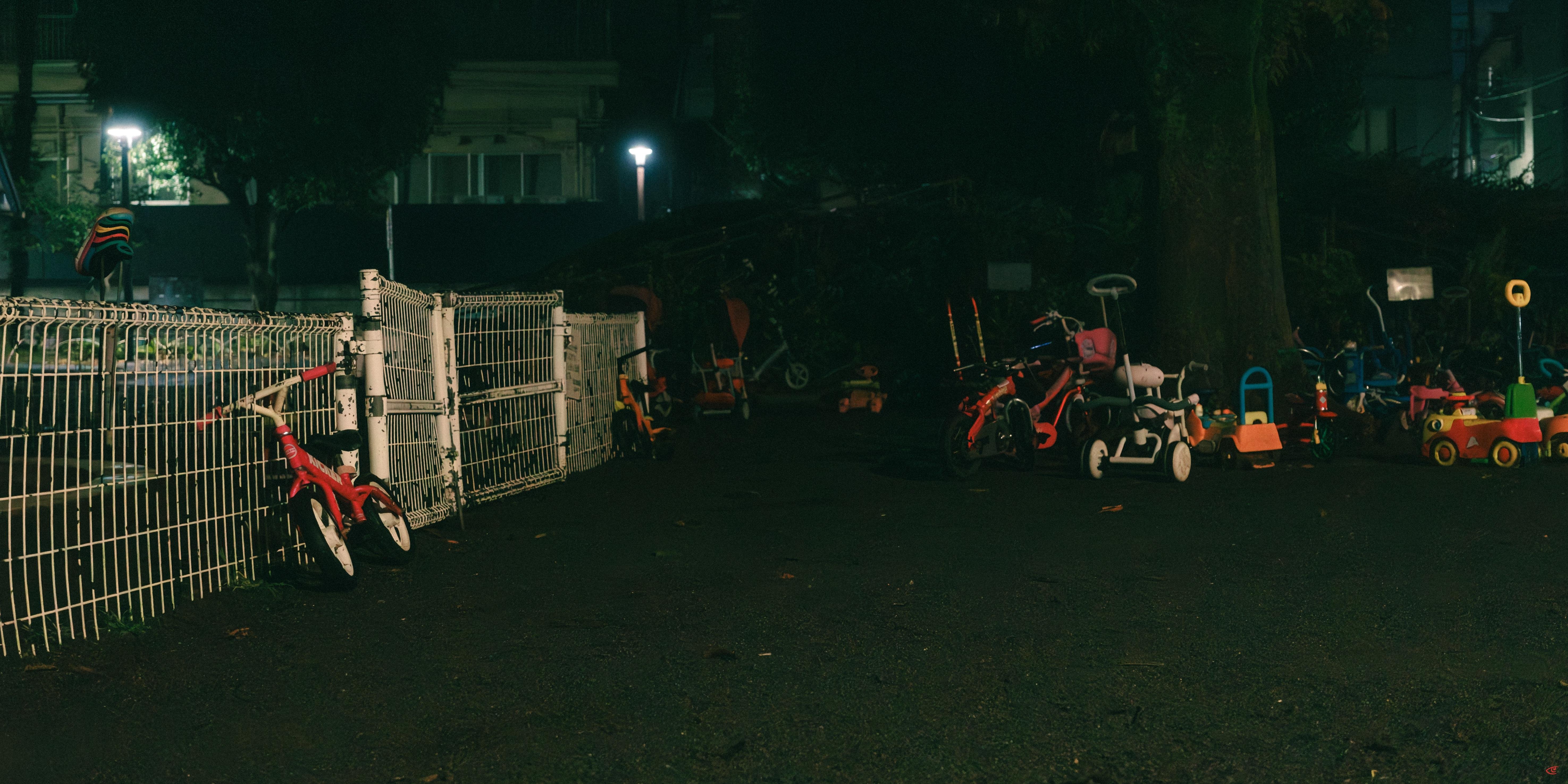 Dimly lit outdoor lot at night with an open metal gate and white fence on the left, a red bicycle leaning against the fence, and several children’s tricycles and ride-on toys scattered along the right side of the asphalt path under streetlights.