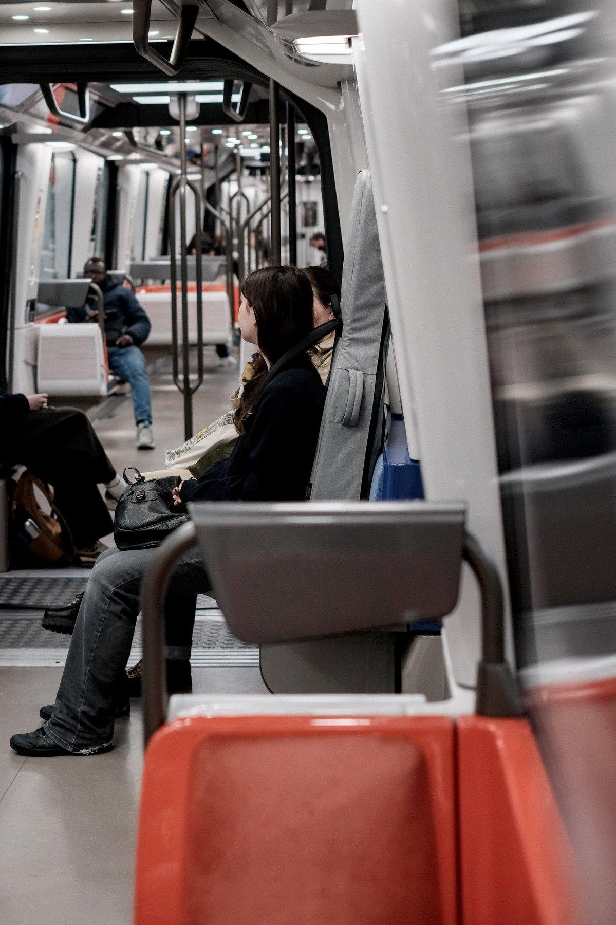 Photo verticale. Dans une rame de métro, une jeune femme en haut noir et jean est assise sur un siège perpendiculaire au sens de la marche, un étui souple de guitare gris dans le dos et son sac à main noir sur les genoux. Elle tourne légèrement la tête à l’opposé de la caméra pour parler à quelqu’un qu’on distingue à peine entre sa tête et l’étui de guitare