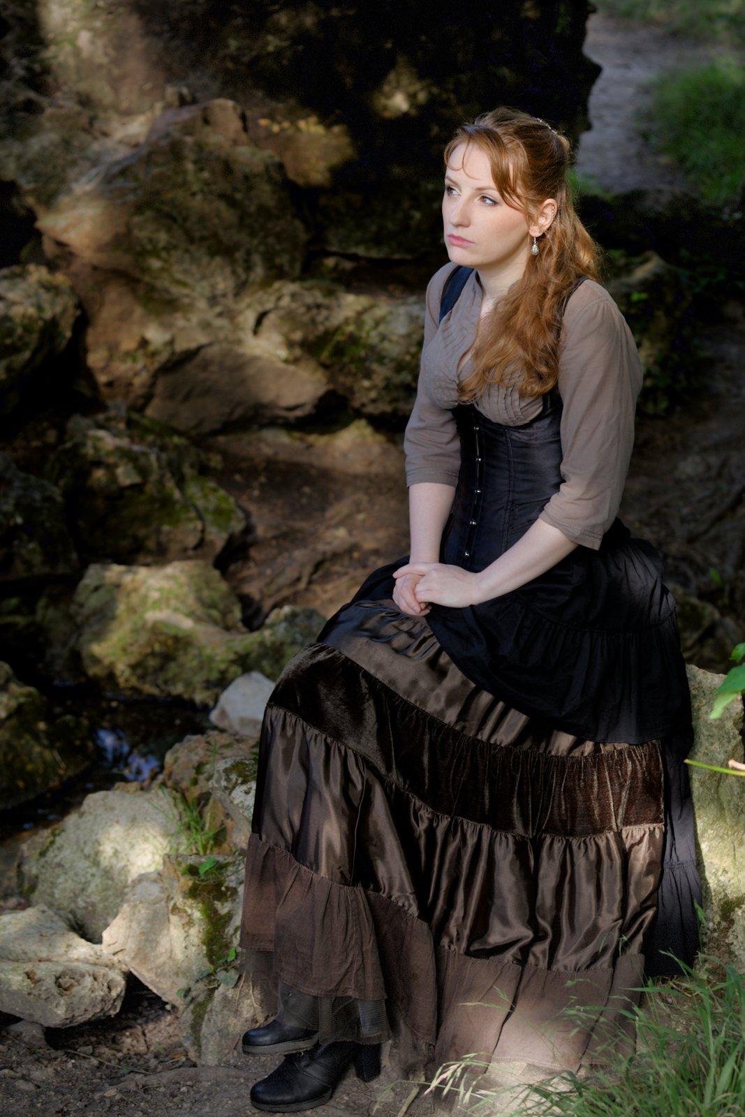 Vertical photo showing a long red hair young woman. wearing a brown shirt with a black corset on top of it, and a long skirt with classy black shoes. She’s sitting on a rock, in the shadow of a tree, with multiple light beam piercing through. She’s looking to some light between the leaves, her hands laying on her crossed leg.