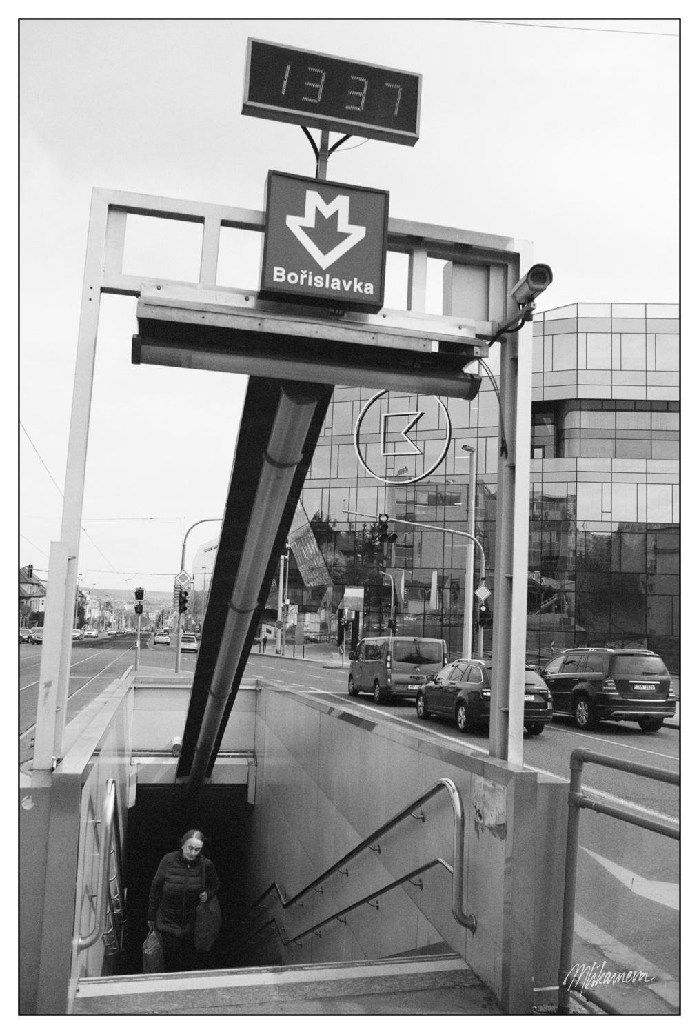 A monochrome photo of a modern looking underground entance with modern looking infrastructure aroun and a person going up the stairs