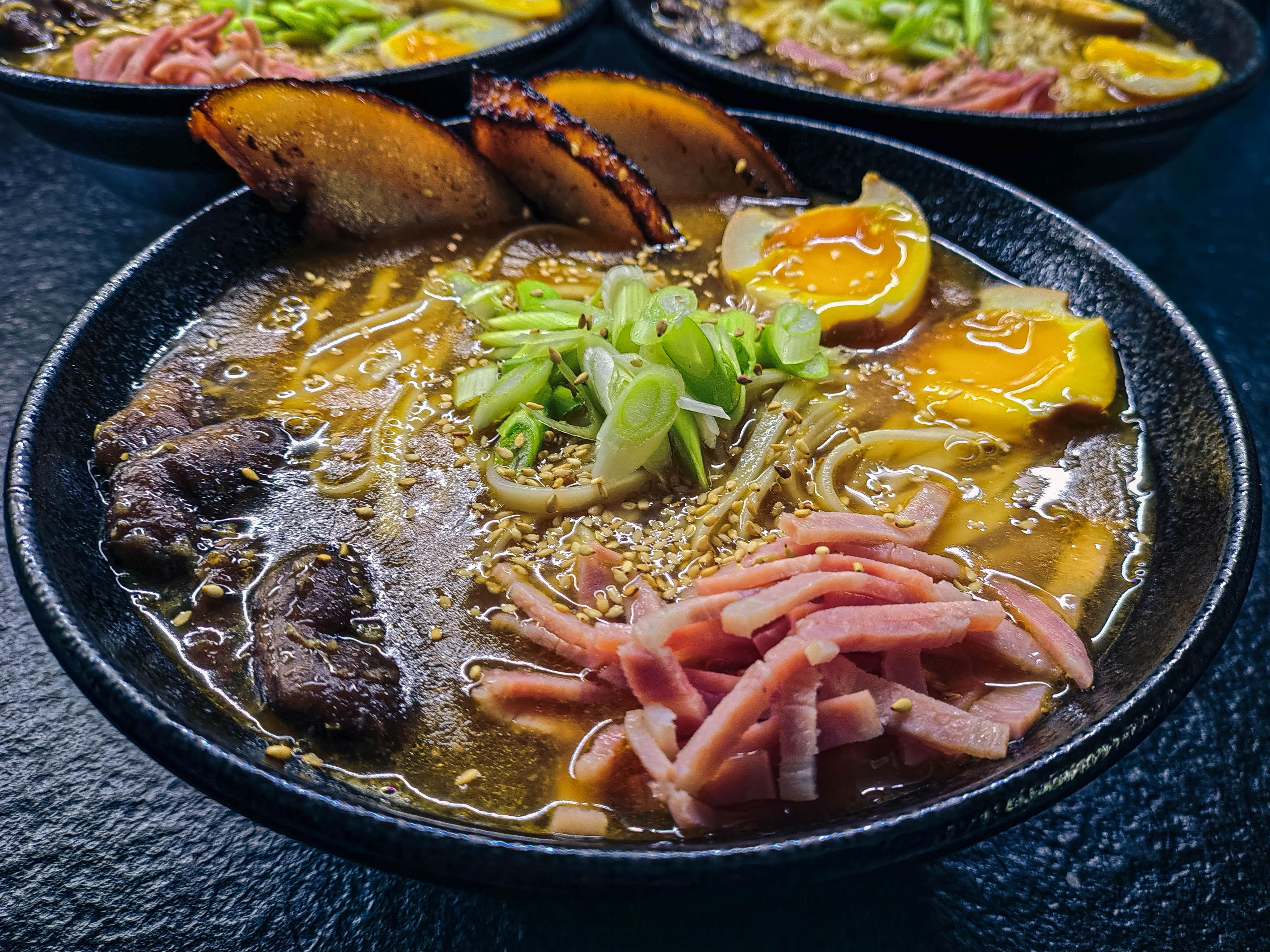 The image shows a bowl of ramen. It includes noodles in broth, topped with sliced green onions, sliced meat (possibly pork), a soft-boiled egg, and a slice of grilled or roasted pork. There are also some sesame seeds sprinkled on top.