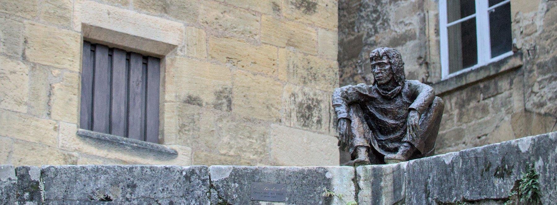 Photo horizontale au ratio très allongé. Une statue d’un homme assis comme un Yakuza au sommet d’un mur