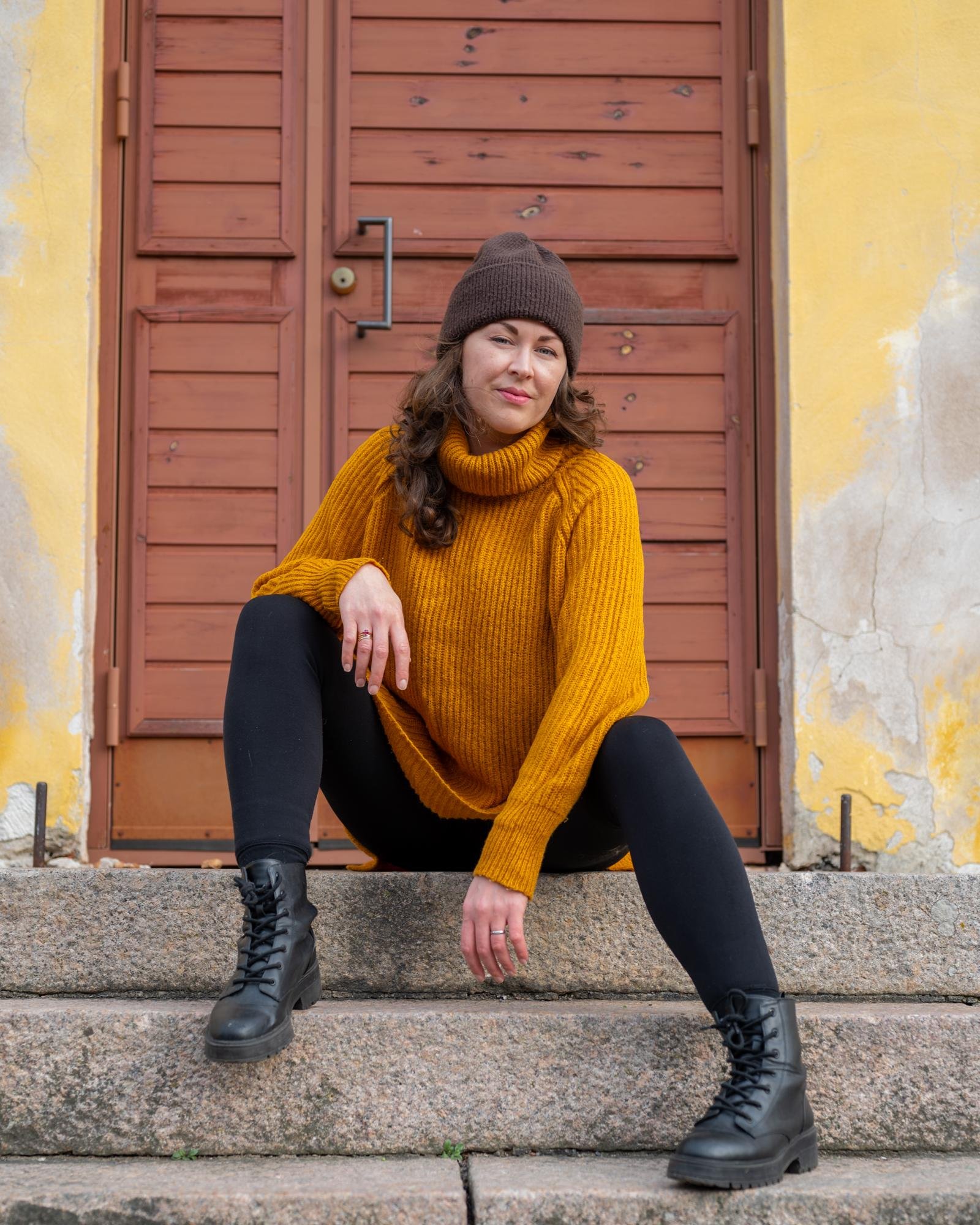 The image shows a person sitting on stone steps. They are wearing a brown beanie, a mustard yellow sweater, black leggings, and black boots. The background features a wooden door and a yellow wall with some peeling paint.