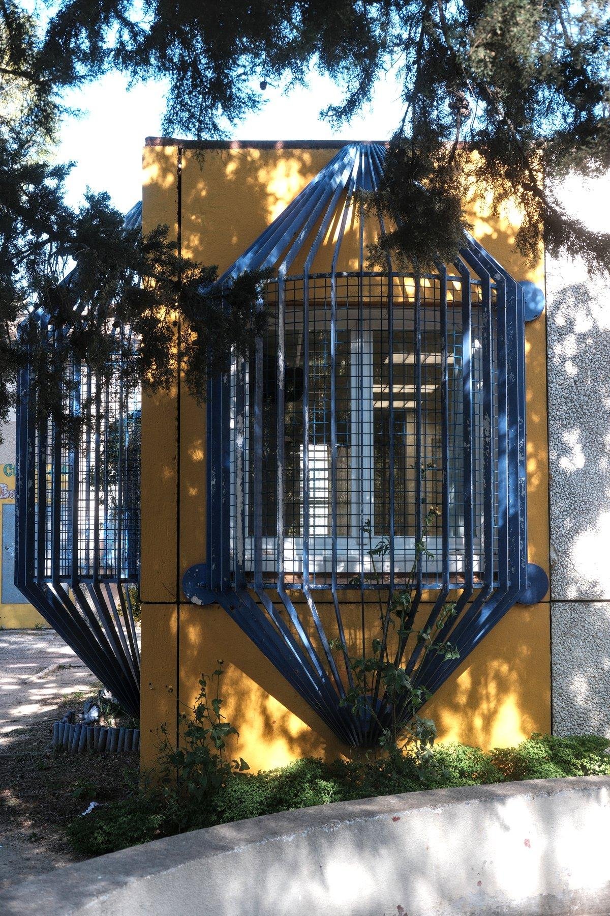 Photo verticale. Une fenêtre dans un mur jaune, protégée par des barreaux bleu qui forme une cage qui se rejoignent en un point unique en haut et un autre en bas. Sur la gauche, on voit une autre cage perpendiculairement, pour une fenêtre qu’on ne voit pas