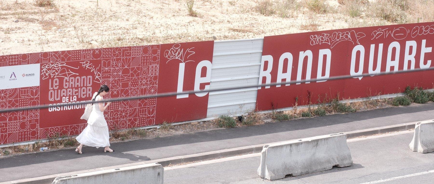 Photo horizontale. Une jeune femme habillée d’une robe blanche et escarpins blancs, marche vers la droite, le long d’une palissage de travaux qui est doté de pancartes rouge aux lettres blanches. Un panneau rouge est manquant, dévoilant le blanc de la palissade en métal, au delà de la barrière, de la végétation jaunie par le soleil qu’on voit assommant