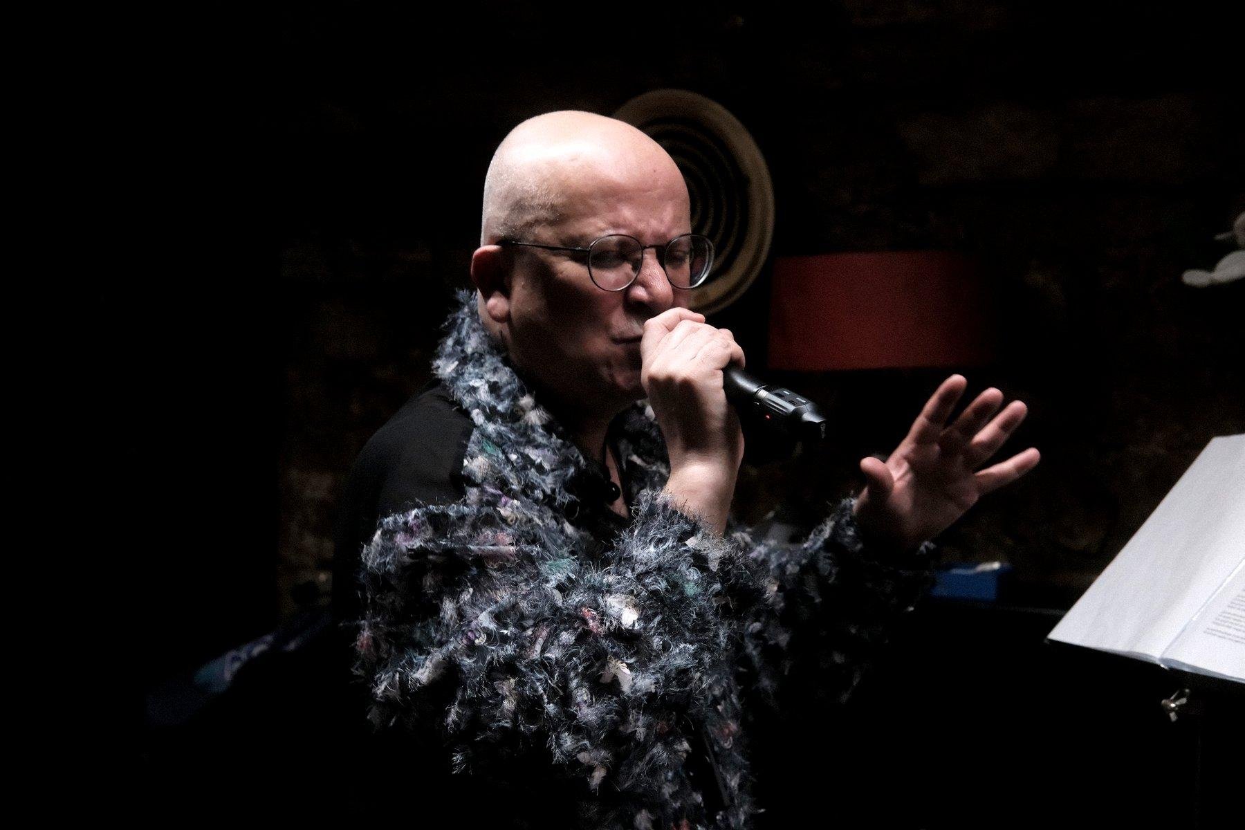 Photo horizontale. Un homme chauve, portant des lunettes et une chemise très décorée, chante avec grande émotion en tenant un micro de la main droite et faisant un geste théâtrale de l’autre main