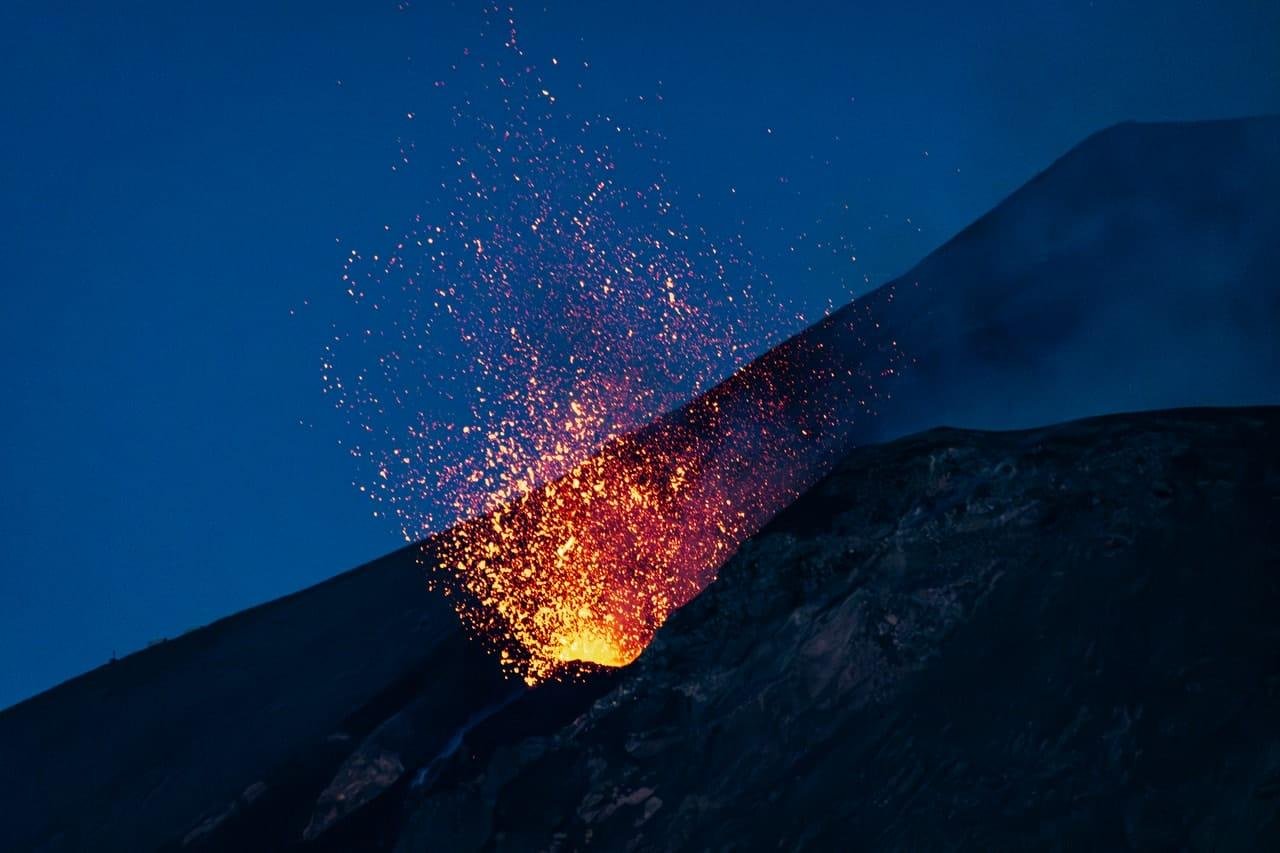 Orange lava fountain and sparks erupting from a volcanic vent on a dark slope at night, with a blue sky in the background.