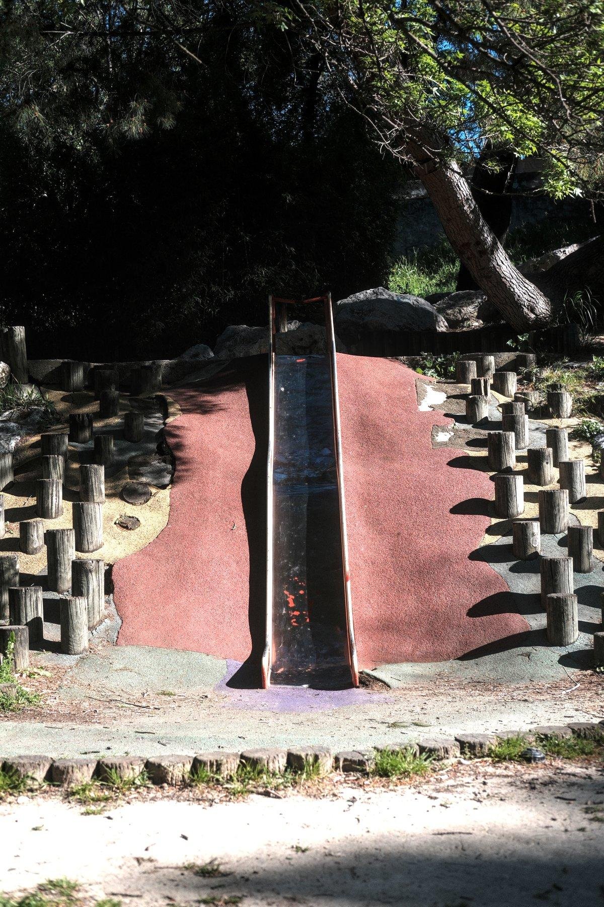 Photo verticale. Un toboggan sur un plan incliné peint en rouge. Il est abîmé avec des éclats dans le revêtement glissant. De part et d’autre du plan rouge, des petits poteaux de bois disposés en quinconce. Devant le toboggan, un sol fait de gravier, au dessus, des arbres.