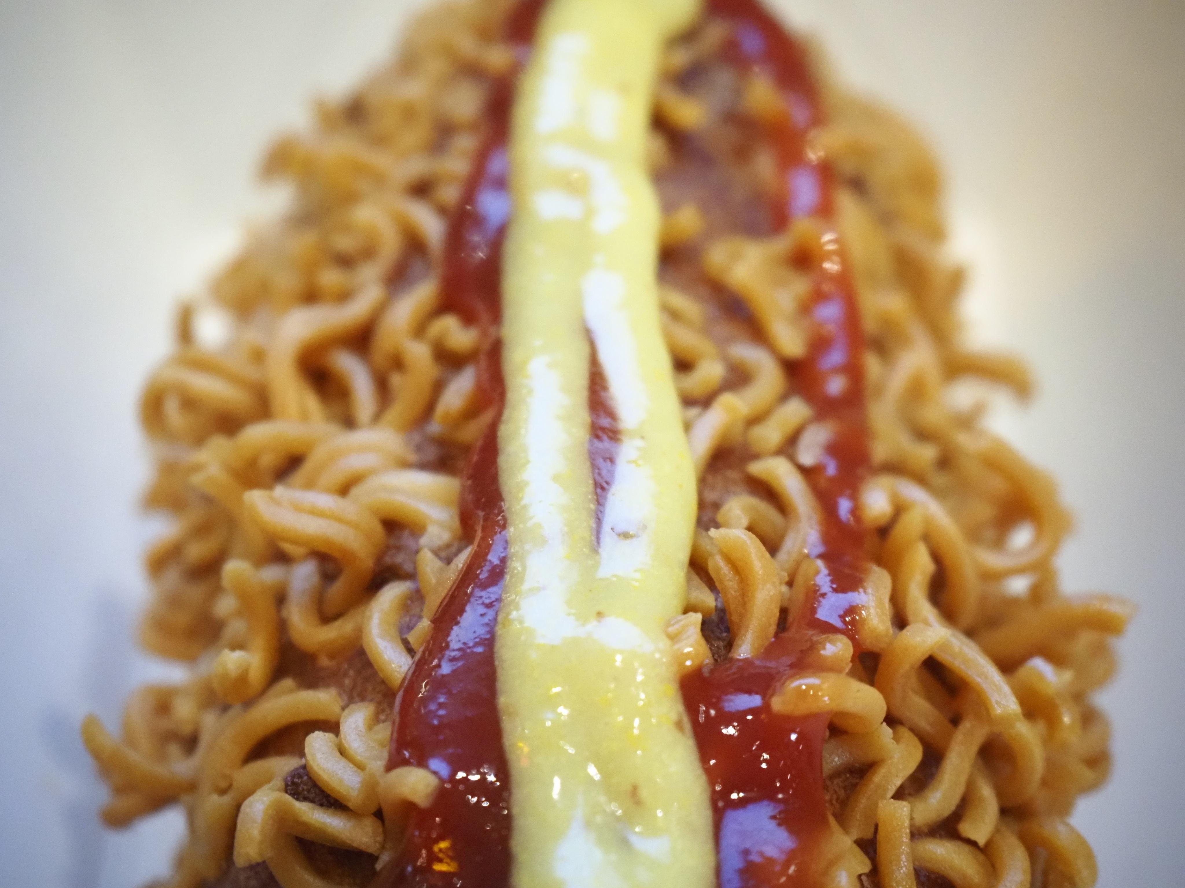 A close-up of a Korean corn dog coated in fried instant ramen noodles. The noodles are golden-brown and cover the entire surface of the dog. Running lengthwise along the top are two parallel lines of sauce — ketchup on either side and a stripe of yellow mustard down the centre. The background is plain white and out of focus.