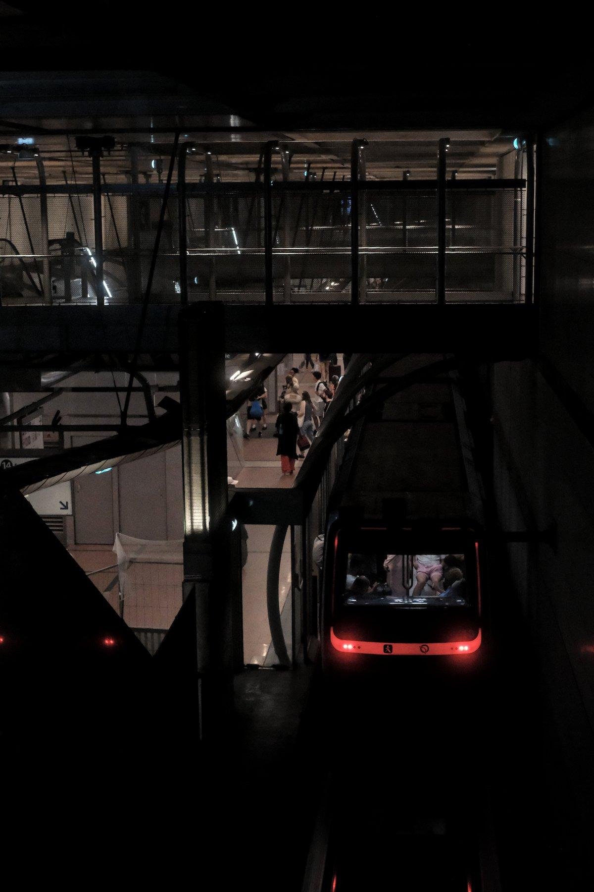 Photo verticale. Vu d’un étage au dessus, à droite, l’arrière d’une rame de métro à l’arrêt avec ses phares rouge, dont on ne devine que les contours dans l’ombre. À gauche, le quai avec des gens. En haut, une passerelle piétonne et sa barrière en verre. En bas à gauche, la paroi d’un escalator