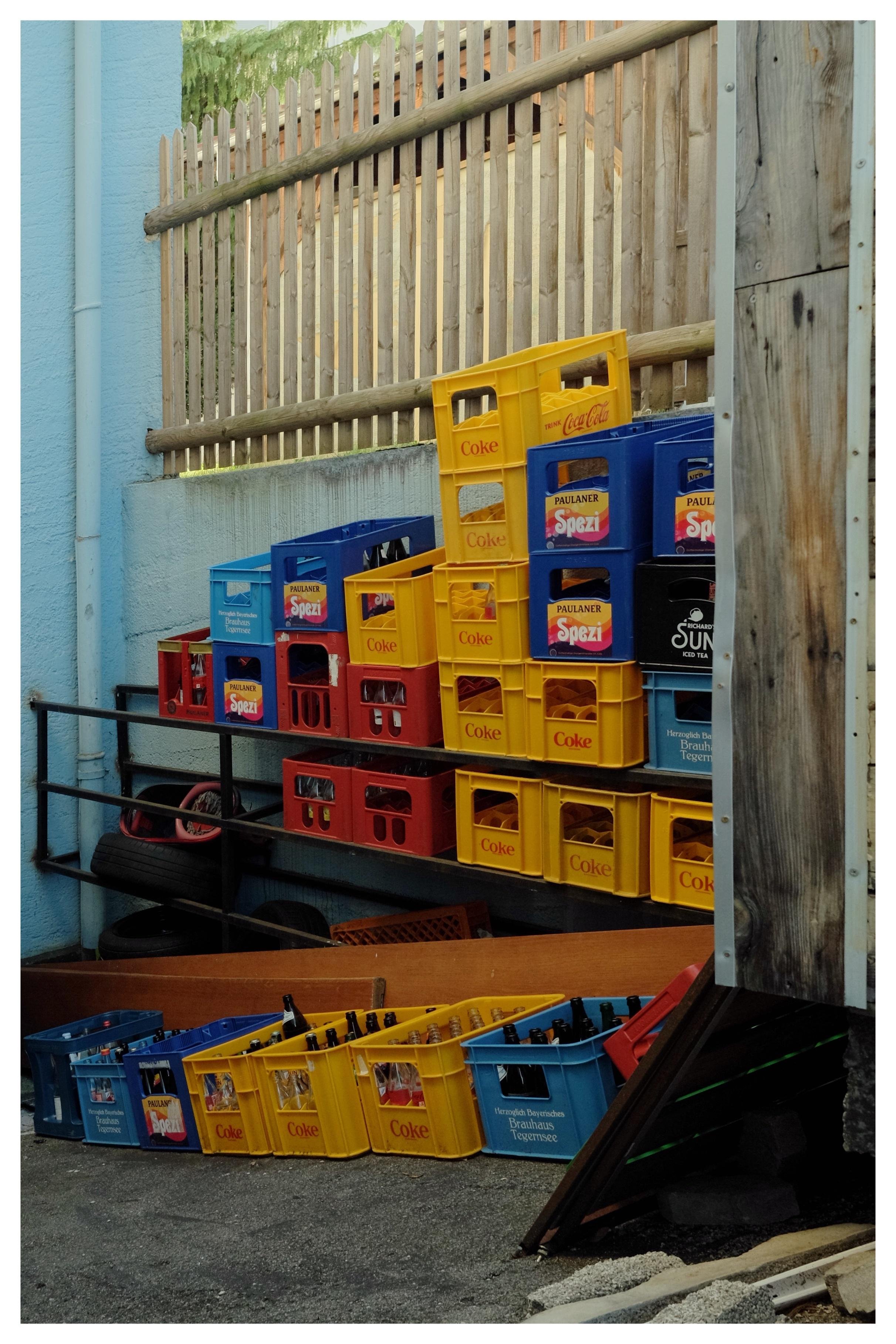 Stacks of plastic beverage crates in yellow, blue, and red filled with glass bottles along a wooden fence and blue wall, with more crates lined on the ground beside a metal railing and tires.