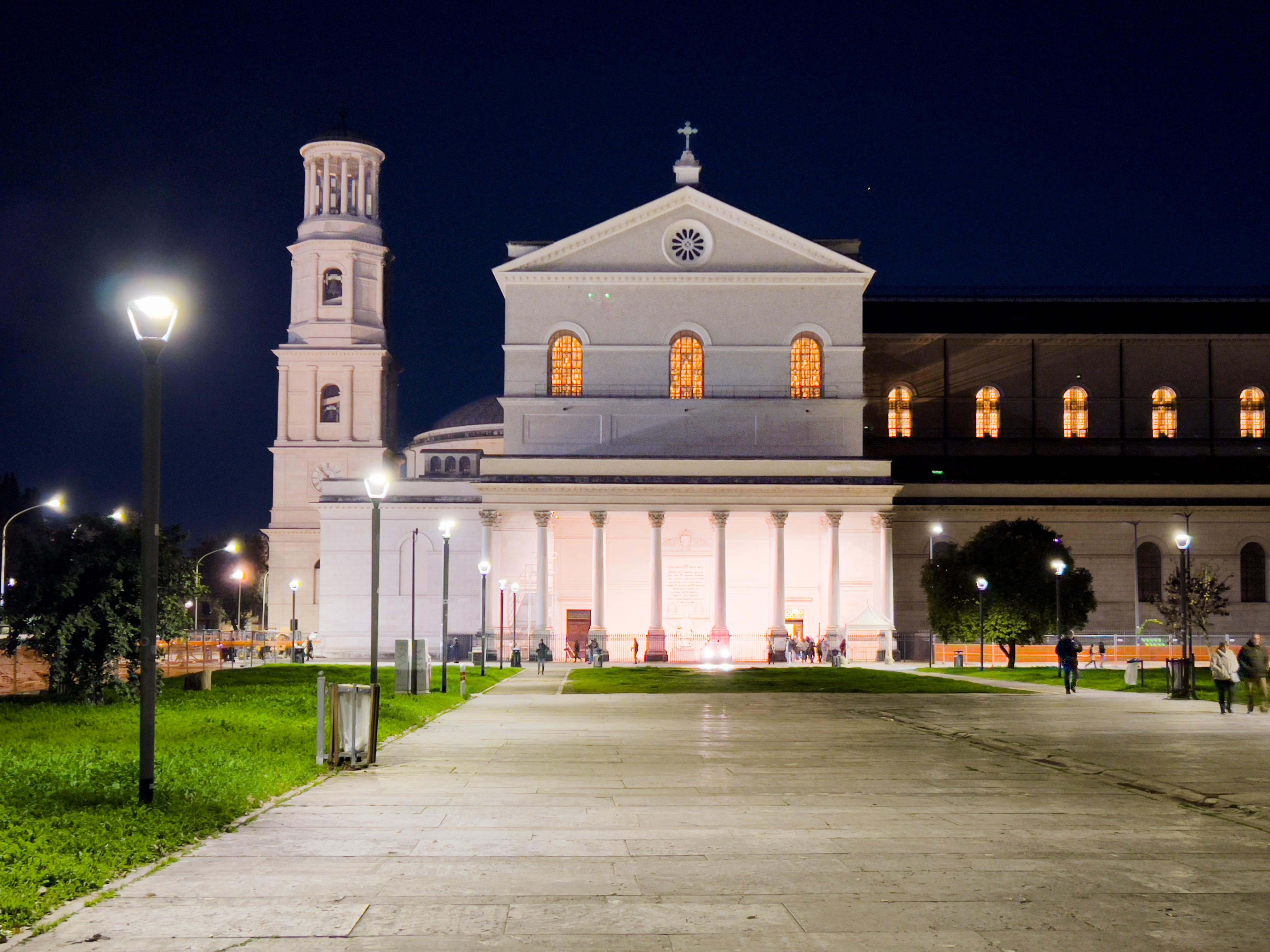 Grande edificio di chiesa illuminato di notte con una facciata prominente caratterizzata da colonne e una croce in cima. A sinistra si trova un campanile e l'area è illuminata da lampioni, con persone che passeggiano intorno. Il cielo è scuro, suggerendo che è notte.