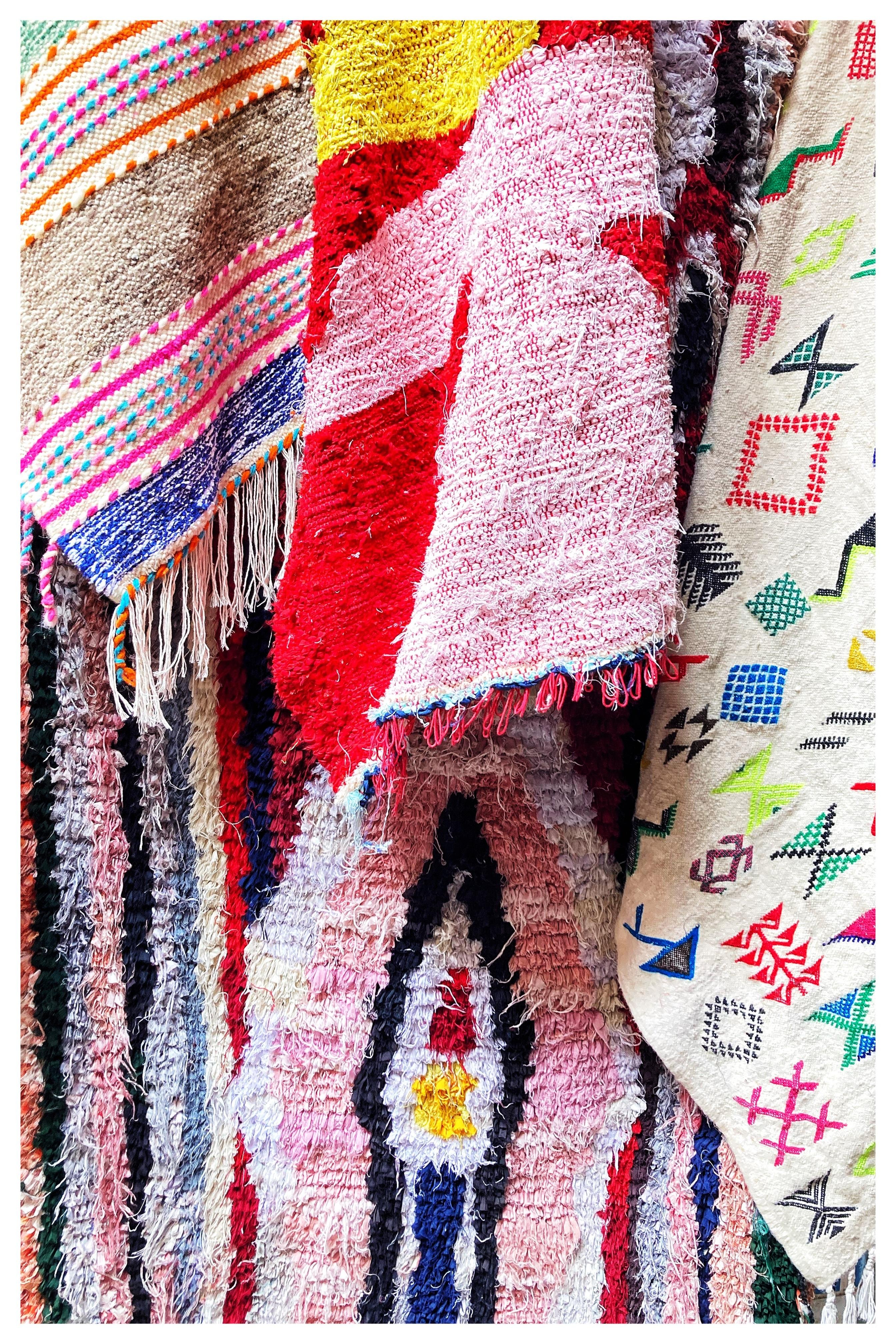 Colorful assortment of textiles featuring various patterns and textures. The fabrics include a striped design with pink, orange, blue, and green accents, a red and pink textured cloth, and another with geometric shapes in multiple colors like green, red, and blue. Fringed edges and diverse woven textures are visible.