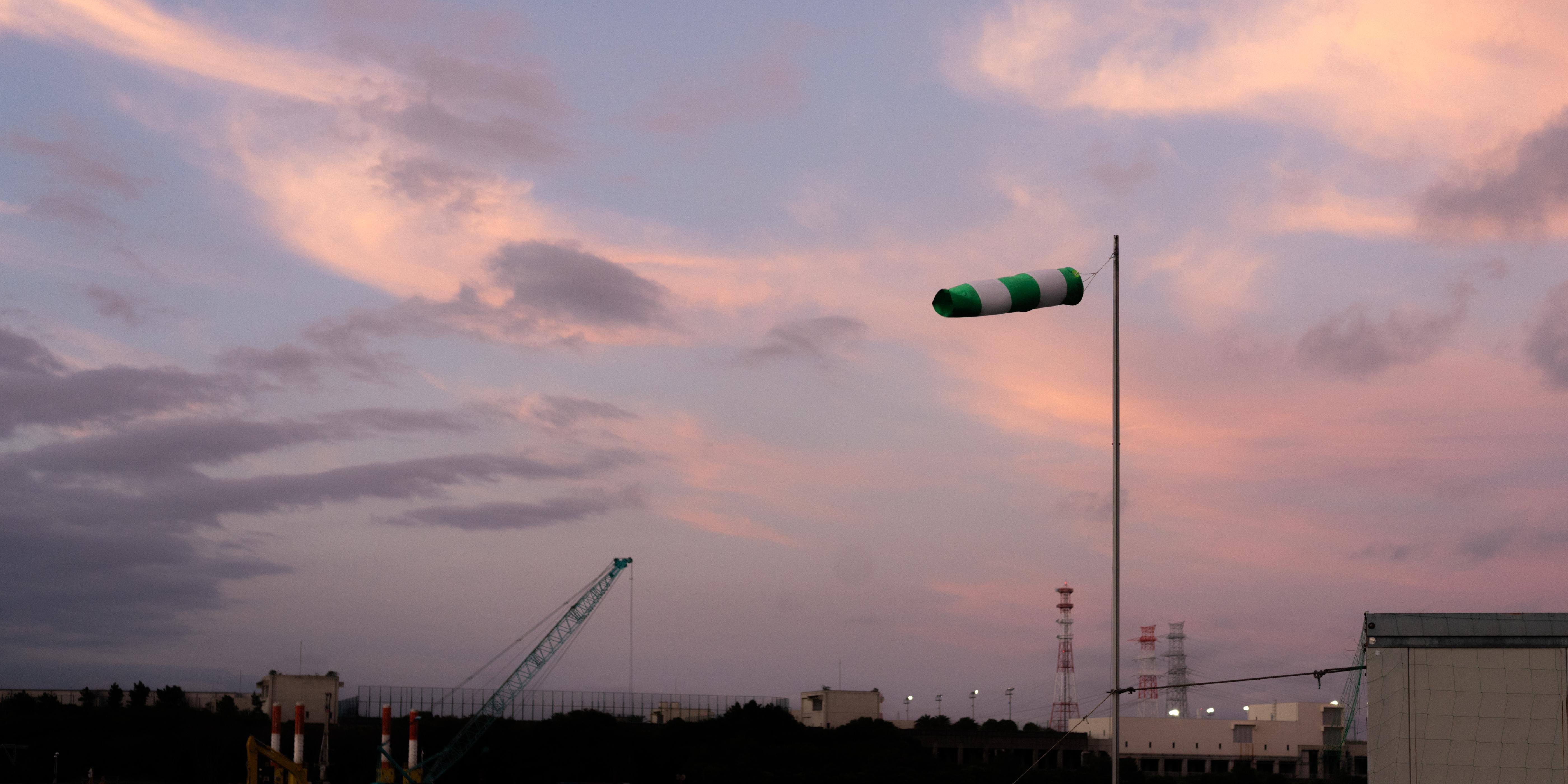 Windsock with green and white stripes mounted on a pole, gently angled due to the wind. Background features a construction crane on the left, several buildings, and radio towers under a dusk sky with pink and purple clouds.