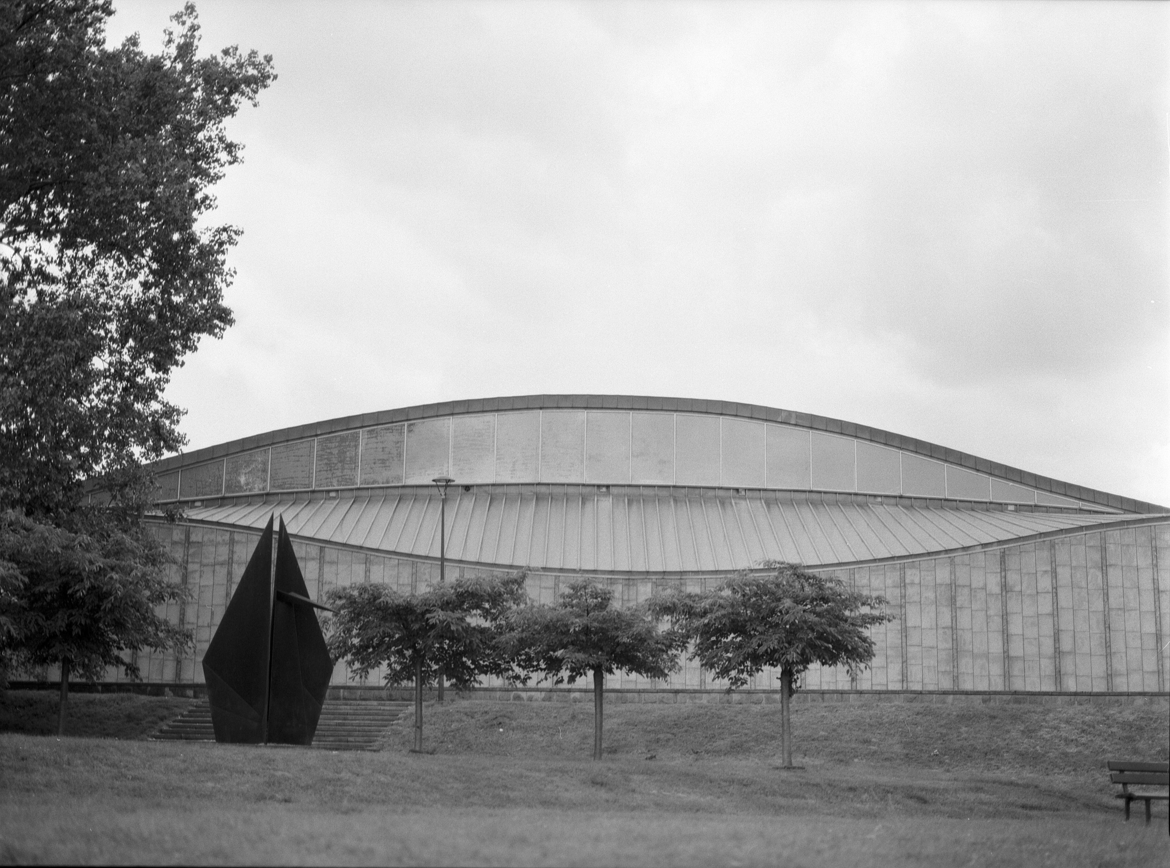 Curved, modern building with a ribbed facade, flanked by trees and a geometric black sculpture. Overcast sky in the background.