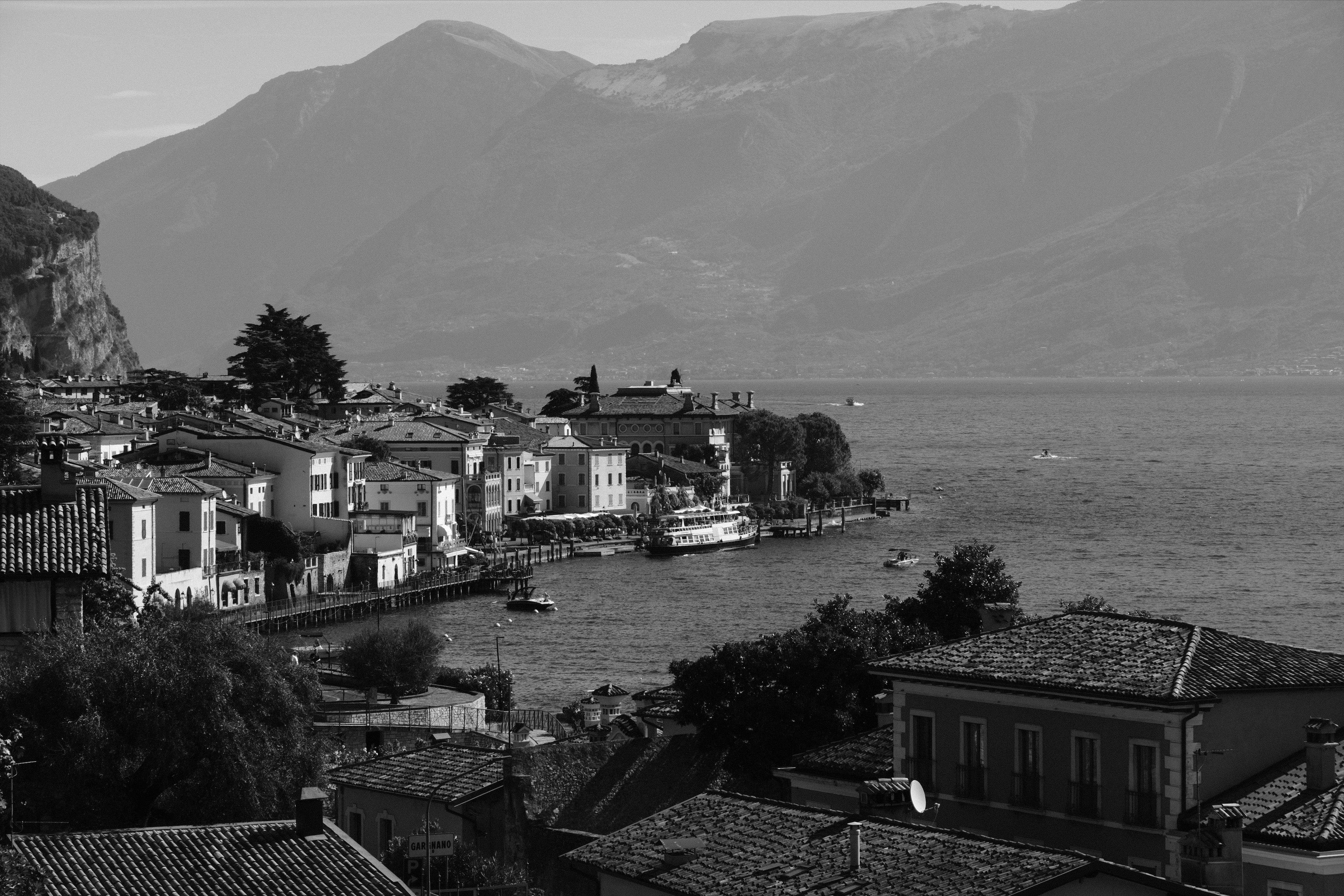 Black and white scenic view of the port and houses along the lake with a ferry docking at the pier. Mediterranean rooftops at the proximal part of the bay in the foreground, the lake and a pale mountain range in the background.