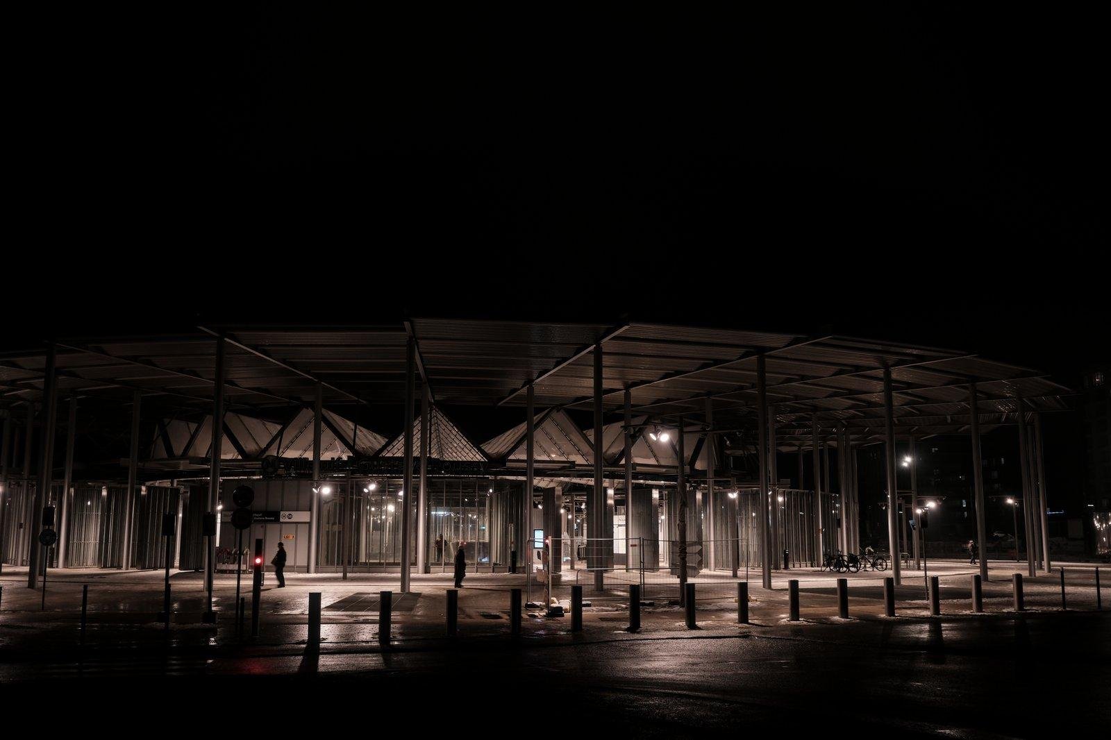 Photo format paysage de nuit, montrant un bâtiment circulaire très ouvert, fait de verre et de métal, avec un grand toit faisant préau et qui rayonne en pleine nuit. Des silhouettes en contre jour passent devant