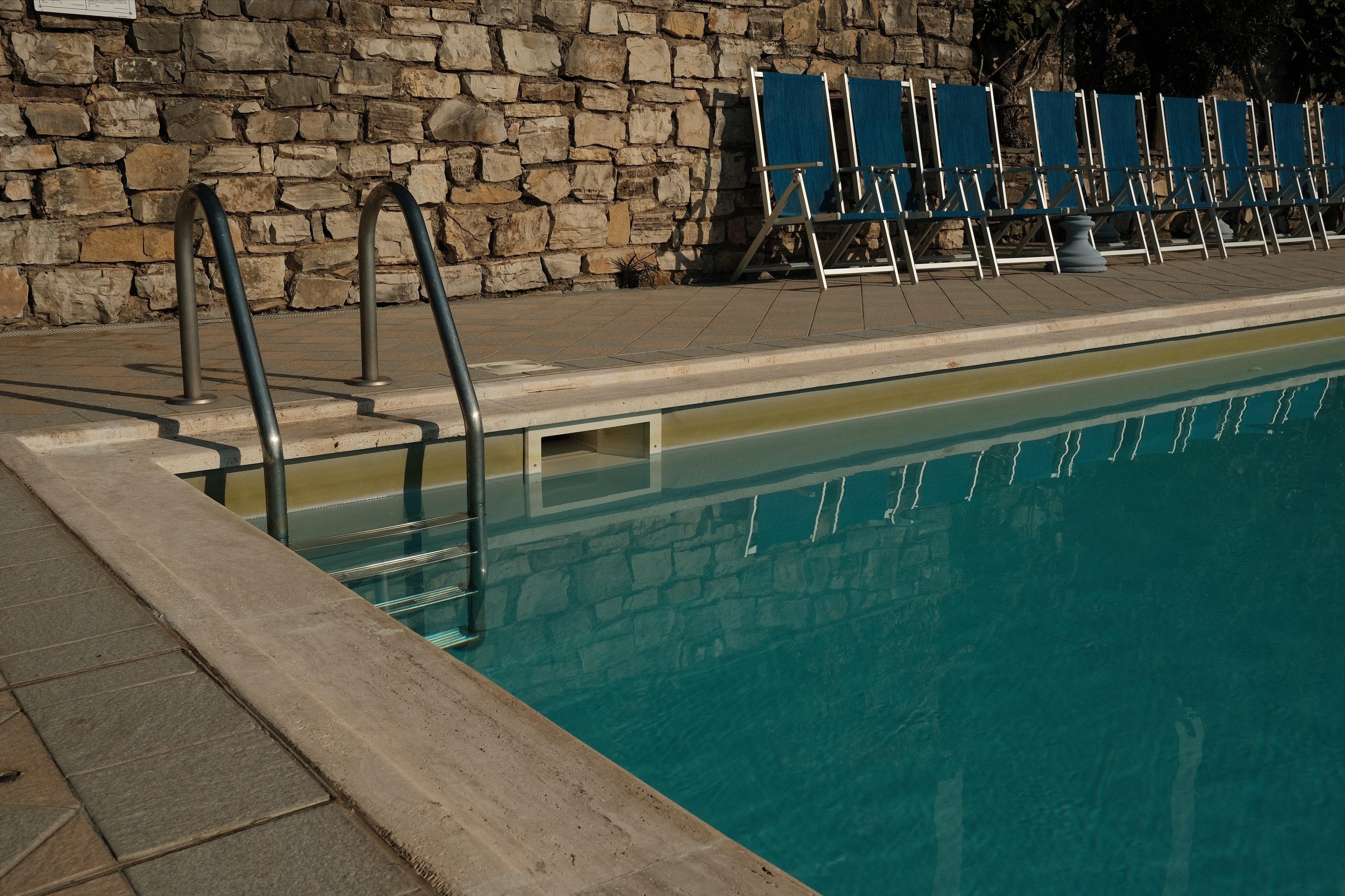 The corner of a swimming pool with a ladder to enter the water, the stone walkway around the pool and blue seats along the walkway to relax.