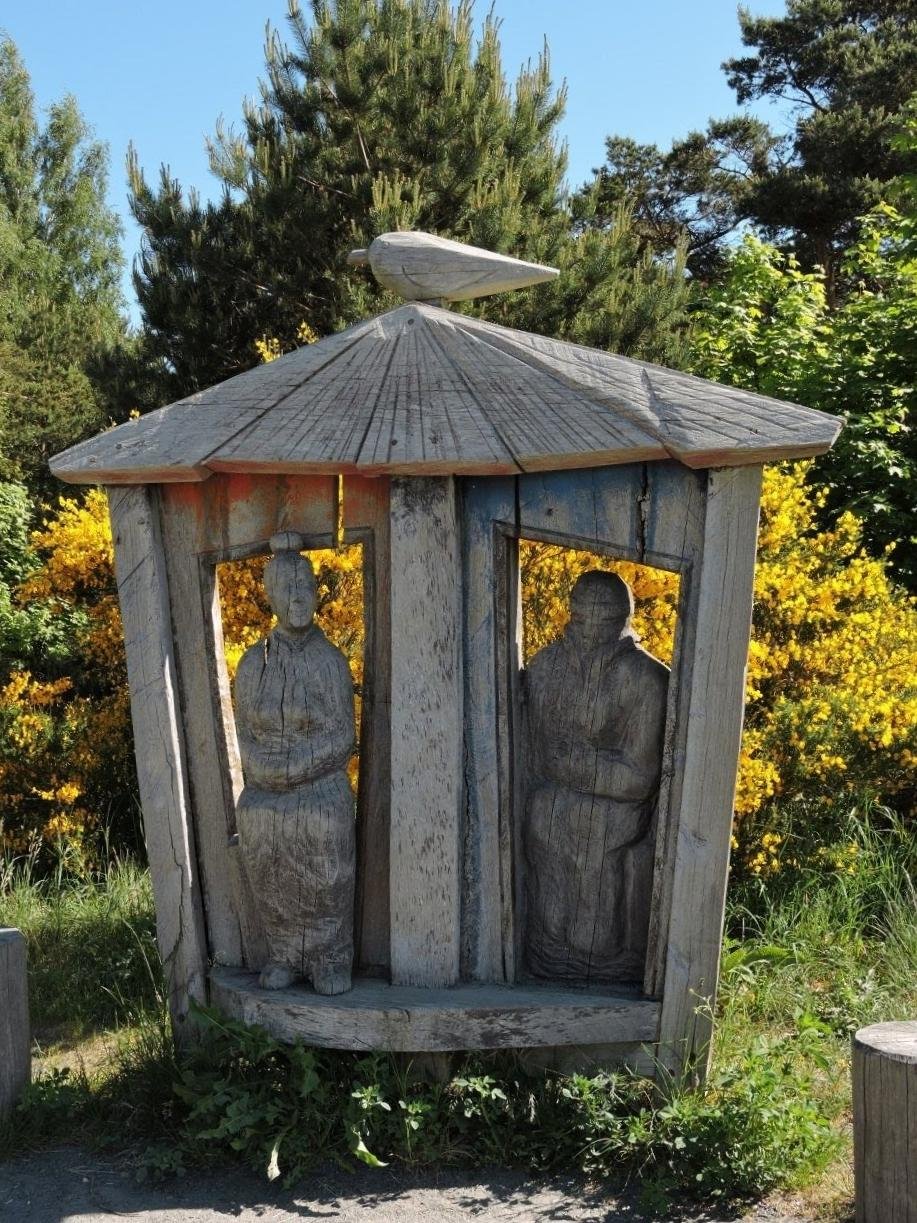 Wooden sculpture of two figures within a hexagonal shelter, topped with a carved bird. Surrounded by lush greenery and yellow flowering bushes under a clear blue sky.