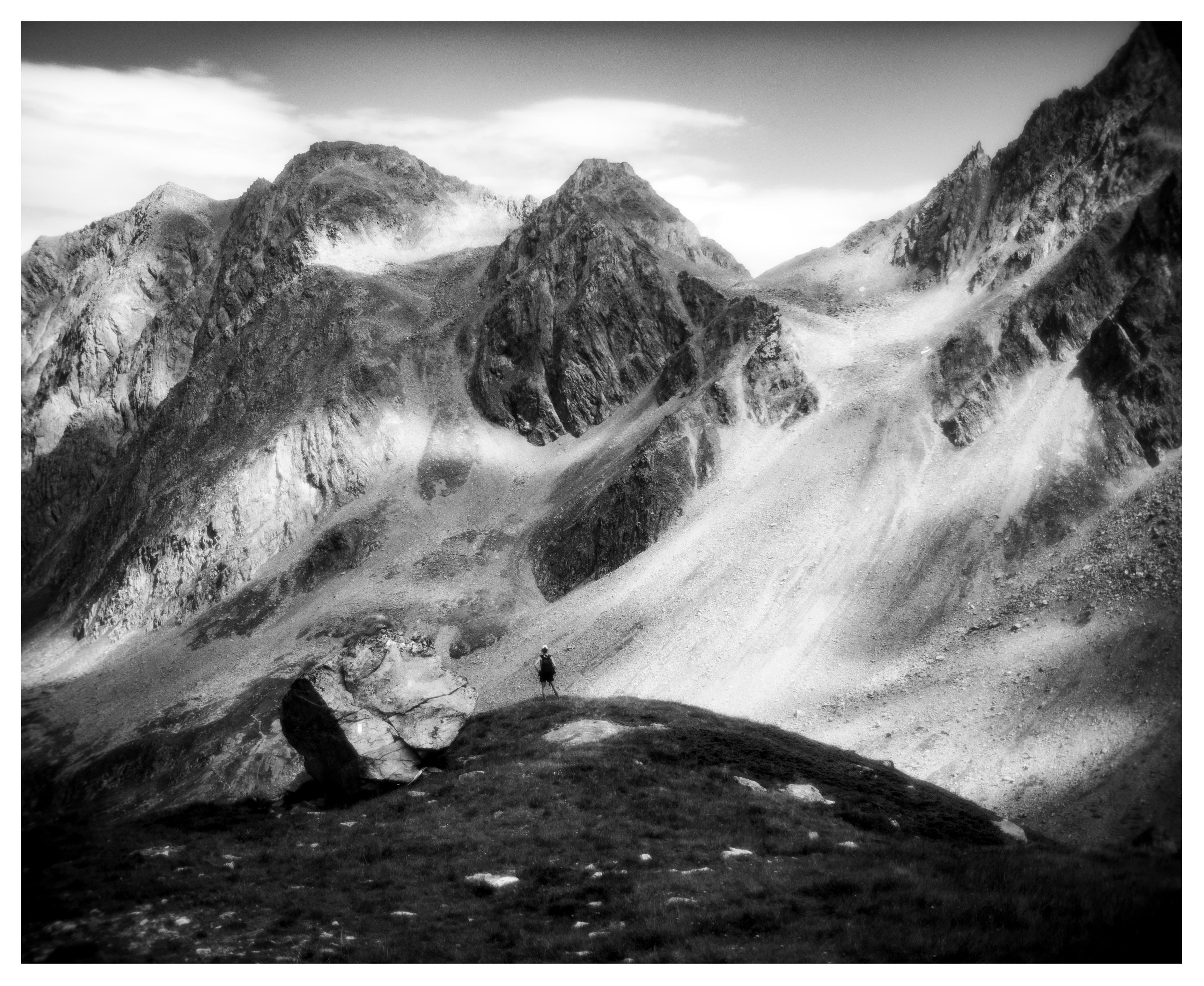 Black-and-white landscape of steep, jagged mountain peaks with snow patches and scree slopes under a cloudy sky; a lone hiker stands on a grassy ridge in the foreground near a large rock.