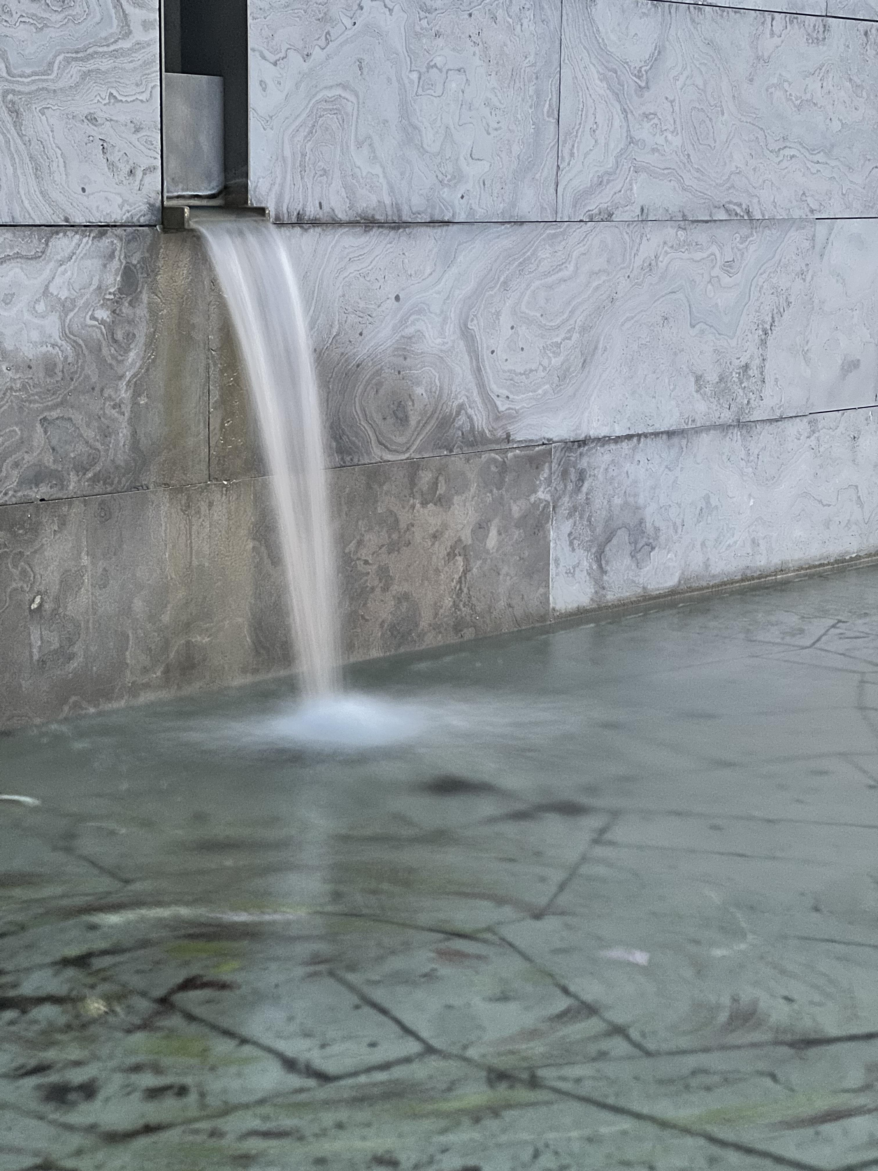 A long-exposure photograph of a stream of water flowing from the outlet and falling into the basin of a fountain feature.