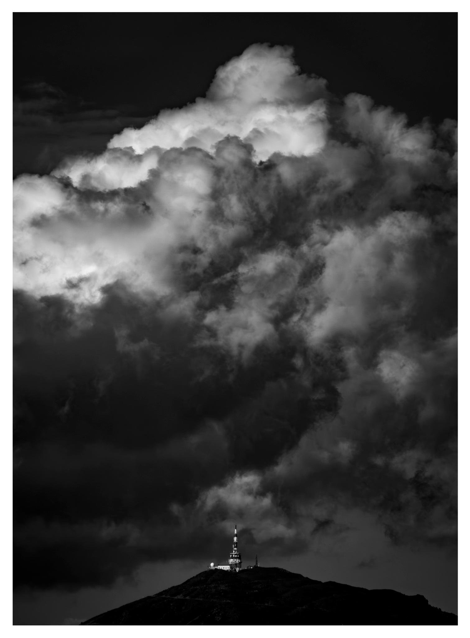 Black-and-white photo of towering, layered storm clouds filling most of the frame, with a small communications tower and buildings silhouetted on a summit.
