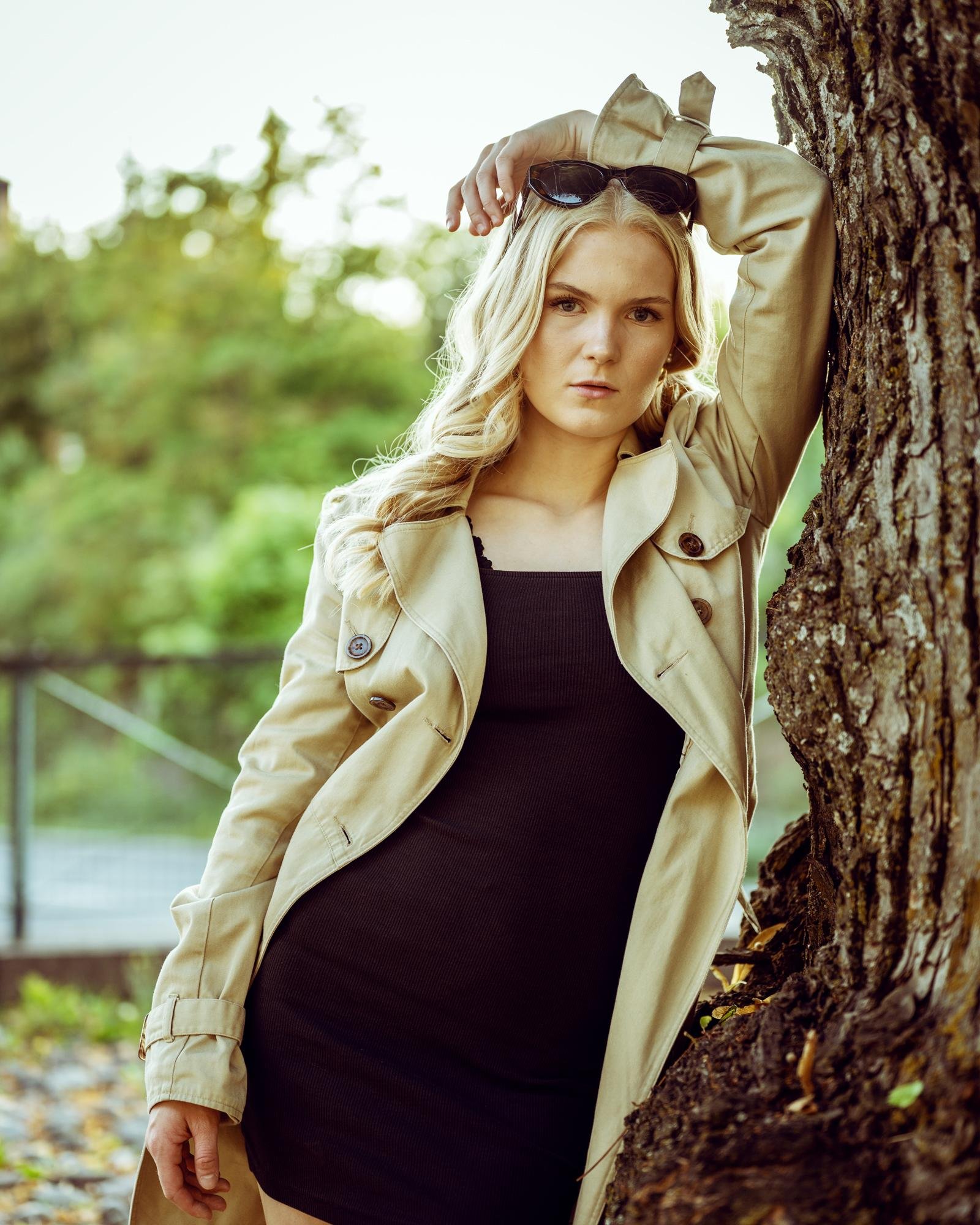 Blonde woman wearing a beige trench coat and black dress, leaning against a tree with one arm raised. Sunglasses rest on her head, and she gazes confidently at the camera. Background features lush greenery, slightly blurred for depth.