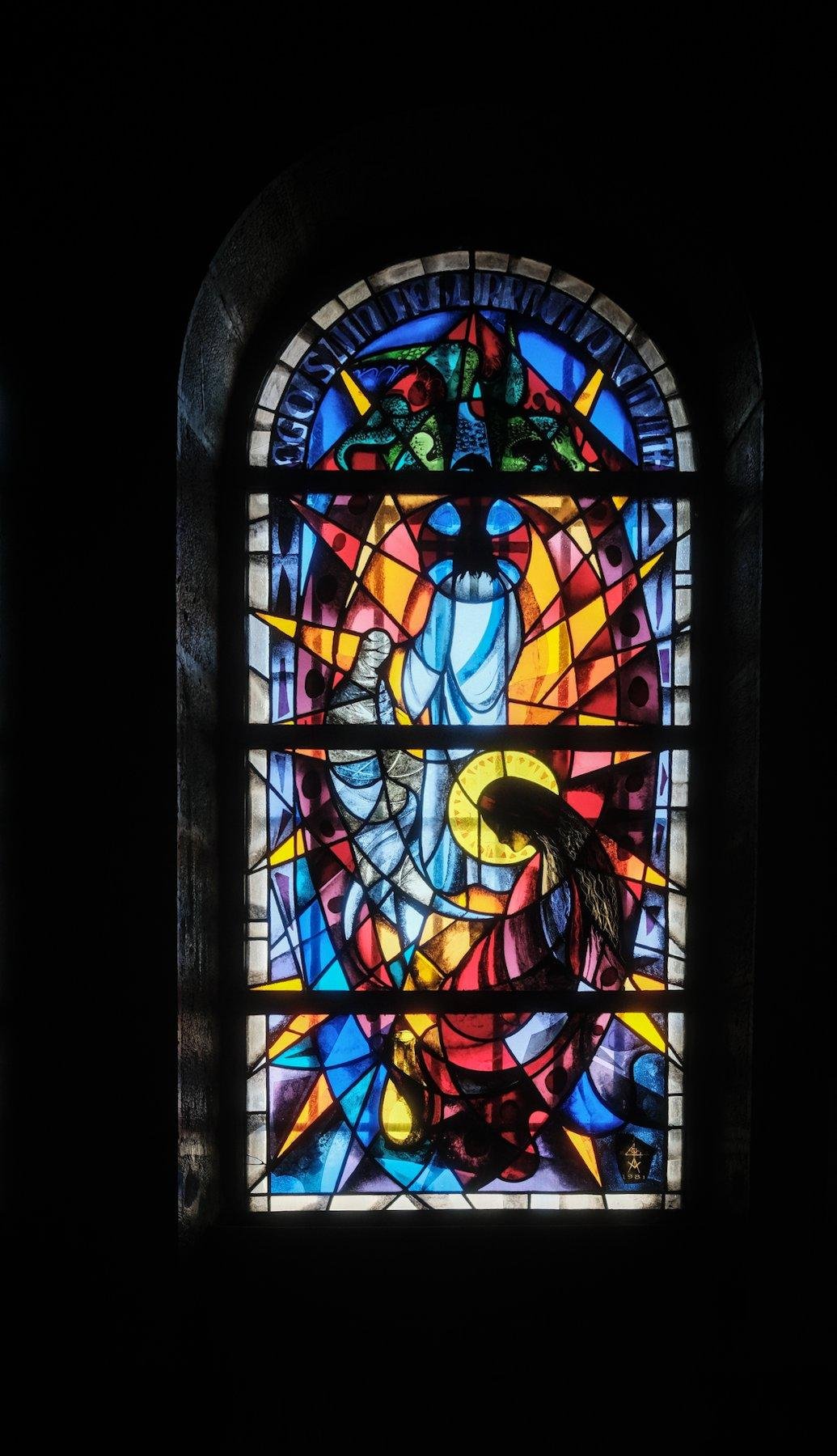 Photo verticale. Des vitraux représentant Marie-Madelaine de profile et Jésus de dos, dans les couleurs bleu, orange et rouge