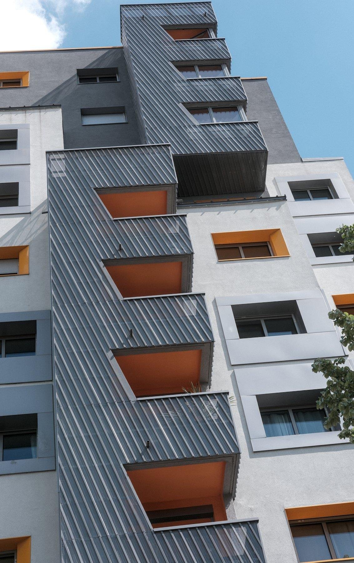 Photo verticale. Contre-Plongée sur un immeuble moderne blanc avec une colonne qui ressort grise en façade et orange sur les plafonds des balcons qui s’y trouvent
