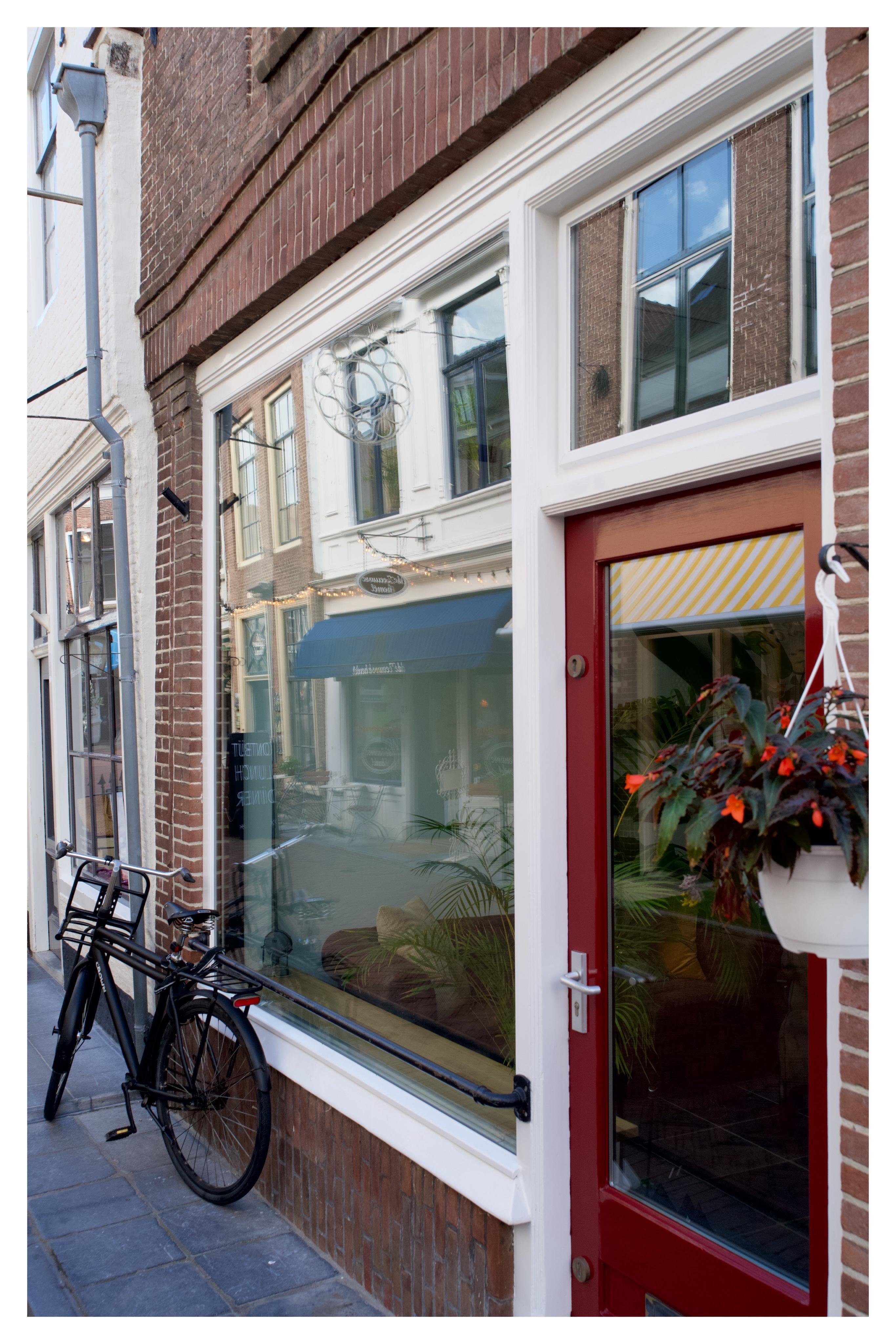 Fassadenansicht einer schmalen Straße mit roter Haustür, großem Schaufenster, Spiegelungen der gegenüberliegenden Häuser und einem schwarzen Fahrrad an der Wand.