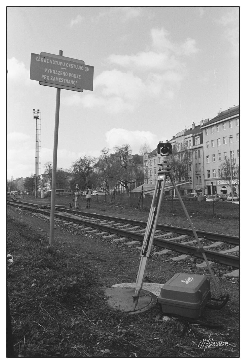 Surveying equipment on a tripod placed beside a railroad track. A nearby signpost reads "Zákaz vstupu cestujících, vyhrazeno pouze pro zaměstnance," indicating restricted access. In the background, there are trees, a person standing further down the tracks, and a row of buildings. A partly cloudy sky is visible above.