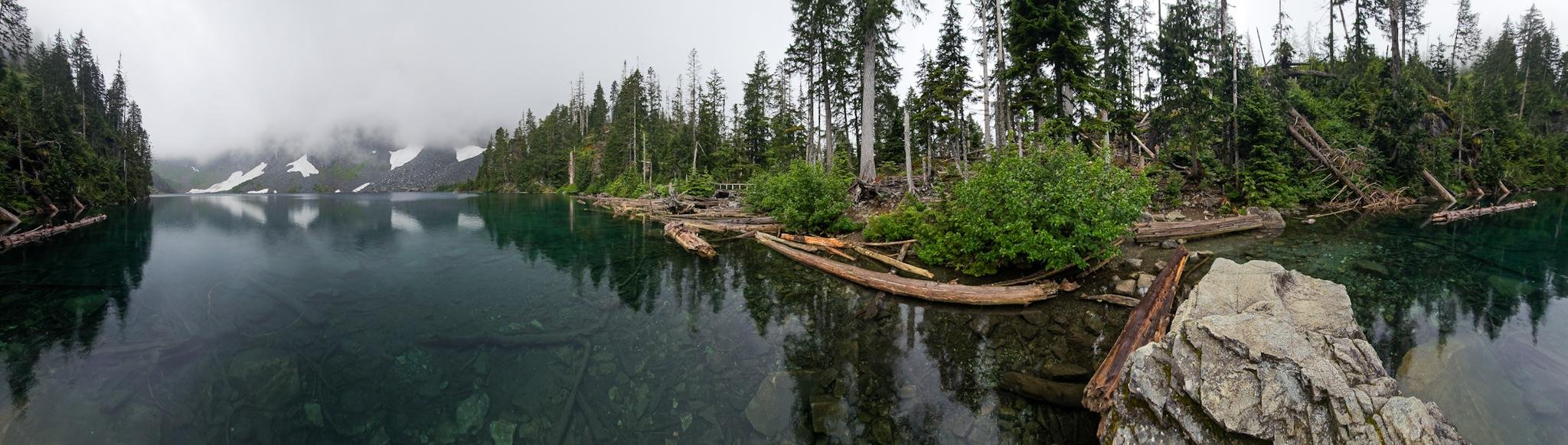 Panoramic view of a calm mountain lake with clear water, a forested shoreline scattered with fallen logs, and a large rock in the right foreground; fog and patches of snow cover the distant mountains under an overcast sky.