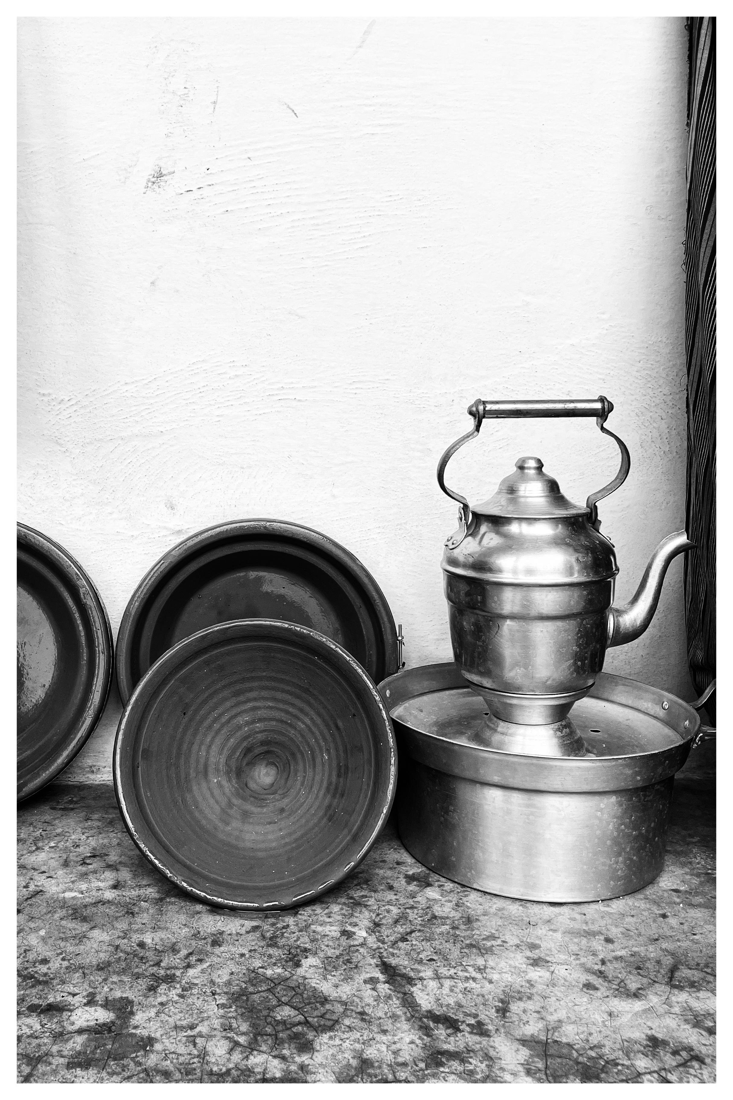 Black-and-white photograph of a metal kettle with a curved spout and handle standing on an upturned basin, with several round clay pots and lids leaning against a plain wall on a worn floor.