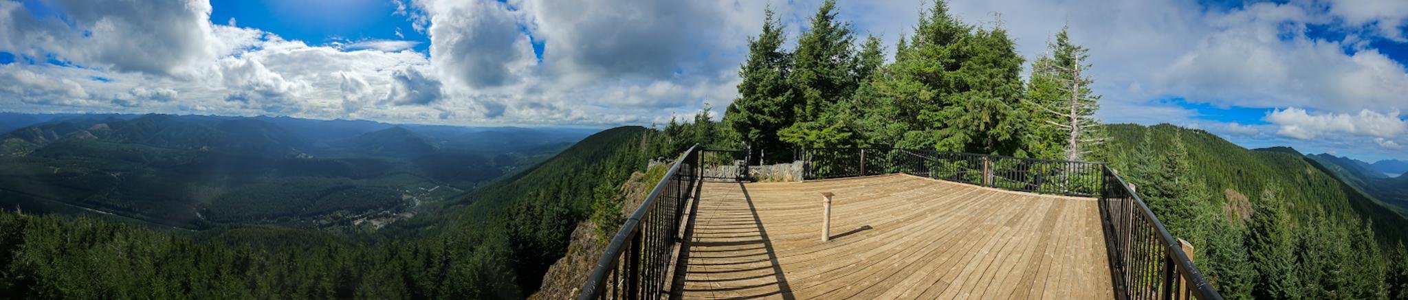 Wide panoramic view from a wooden lookout deck with black metal railings, overlooking forested mountain ridges and a distant valley under a partly cloudy blue sky.
