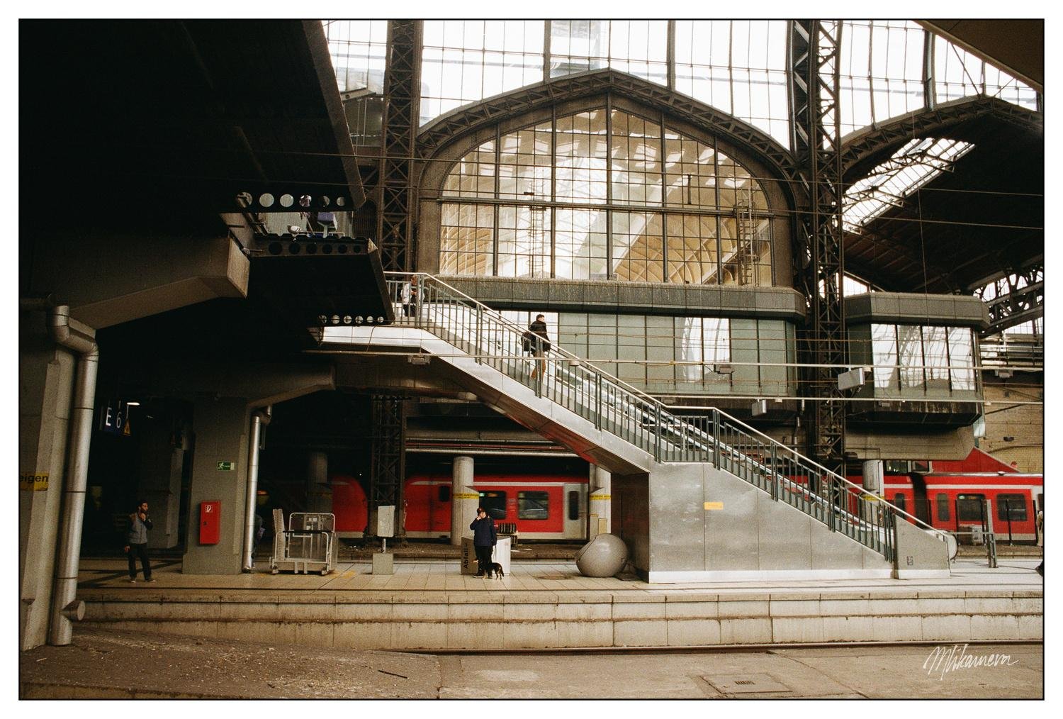 Interior of a train station with a large arched glass roof and steel beams. A staircase leads to an upper level walkway. Two red trains are partially visible on the tracks. People are walking and some are standing near the stairs. A fire extinguisher and equipment are positioned on the platform.