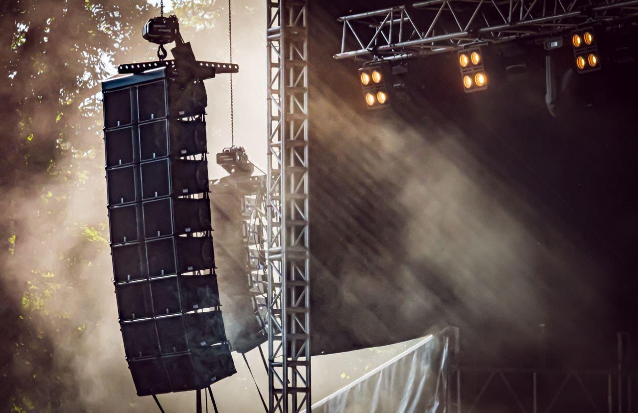 Stage setup with large hanging speakers and spotlights against a backdrop of mist and sunlight. Steel trusses support the equipment, and beams of light cut through the haze, creating a dramatic effect.
