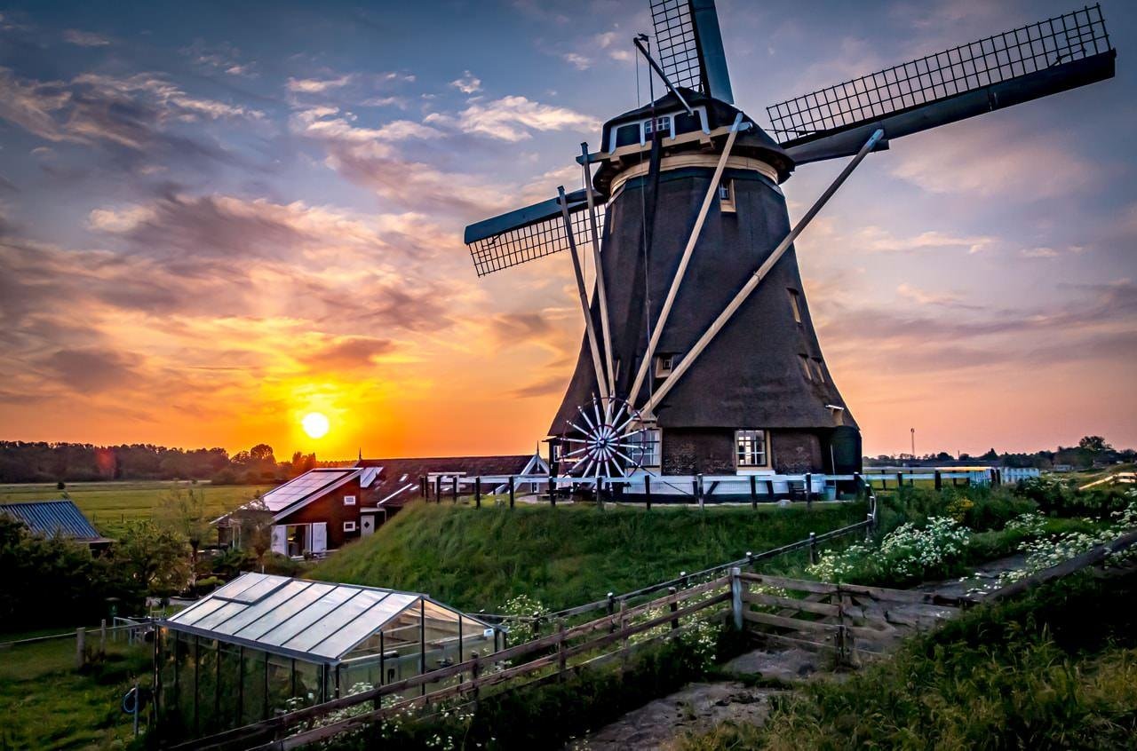 The image depicts a traditional windmill set against a scenic backdrop of a sunset. The sky is vibrant with colors, and there are some small buildings and vegetation surrounding the windmill. This kind of landscape is often associated with regions like the Netherlands.