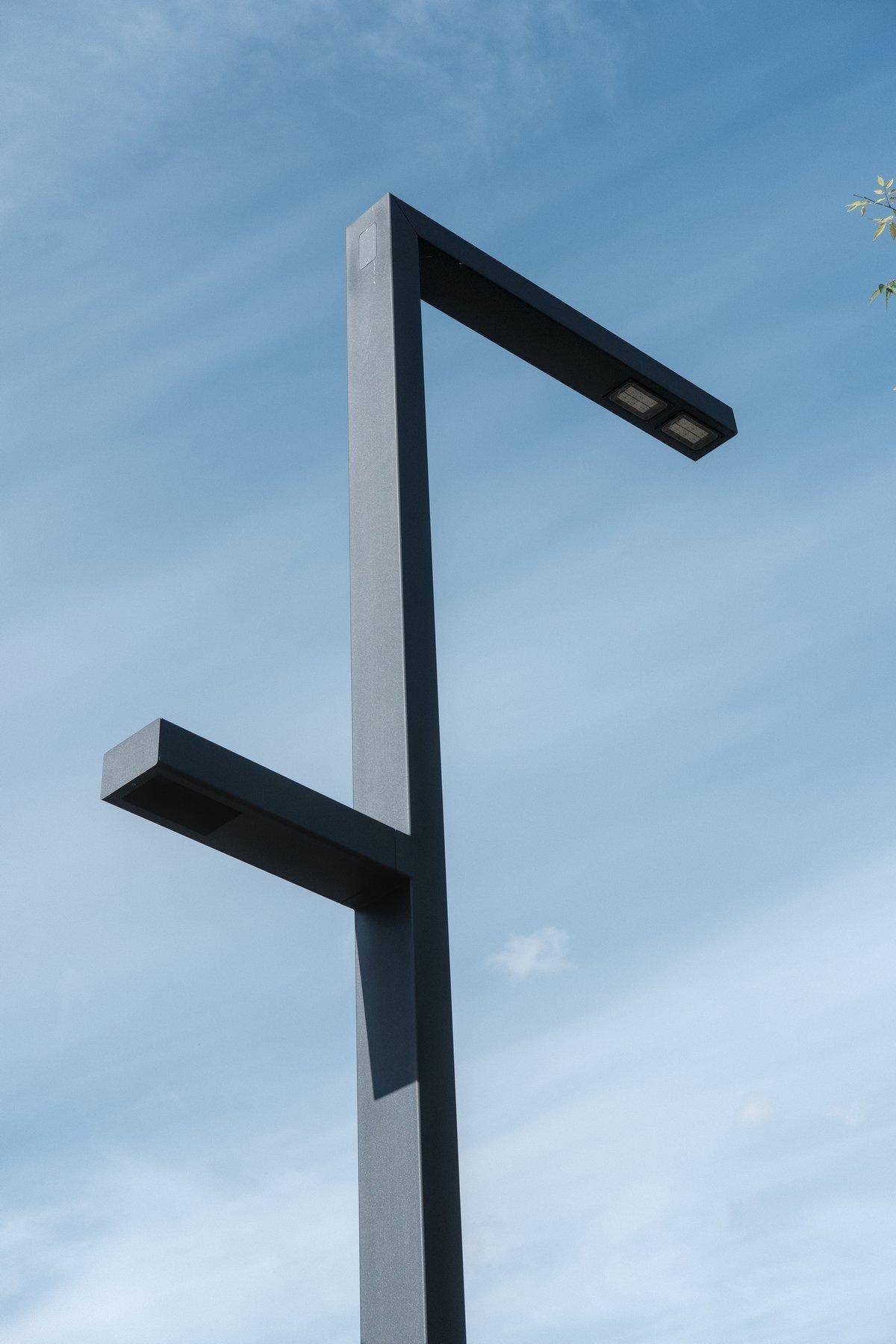 Photo verticale. Un lampadaire gris sombre, très moderne, un poteau rectangulaire se terminant en angle droit sur la route. De l’autre côté, une plus courte protubérance en angle droit aussi, pour le troitoir. Le tout sur fond de ciel bleu avec des voiles de nuages