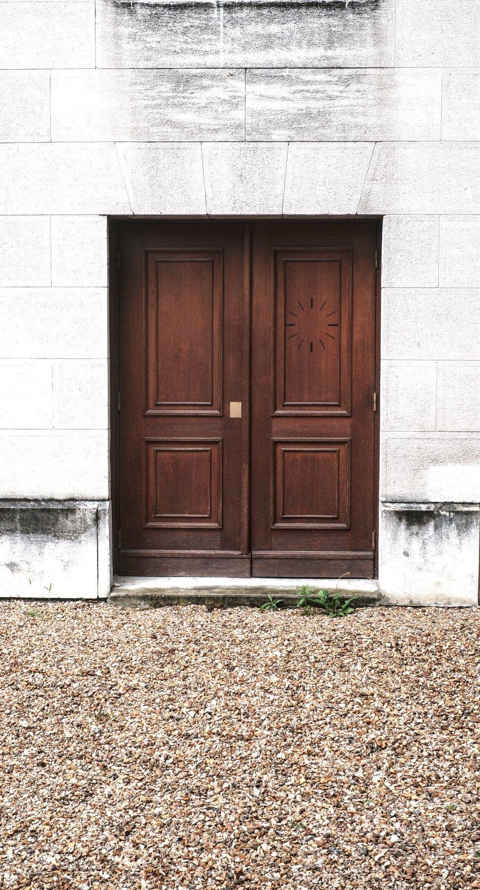 Photo verticale. Une double porte en bois sans grand chose de particulier si ce n’est sur la porte de droite, une espèce d’horloge mais sans aiguille est gravée. Devant la porte, un sol fait de gravier