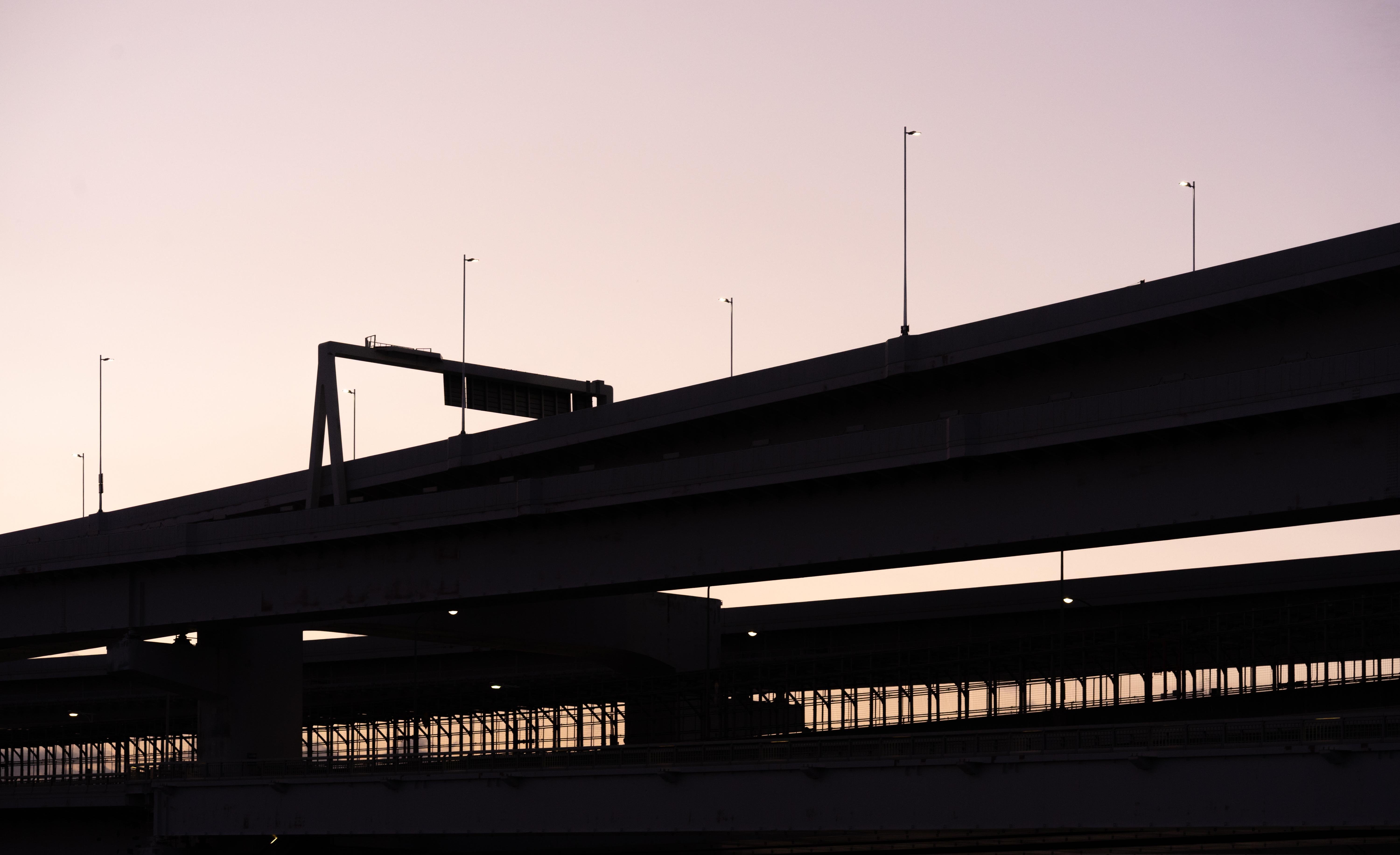 Three layered highway overpasses silhouetted against a gradient sky transitioning from pale pink to deeper purple. Thin light poles line the highways, and a gantry is visible on the uppermost level. The scene is free of vehicles and people.