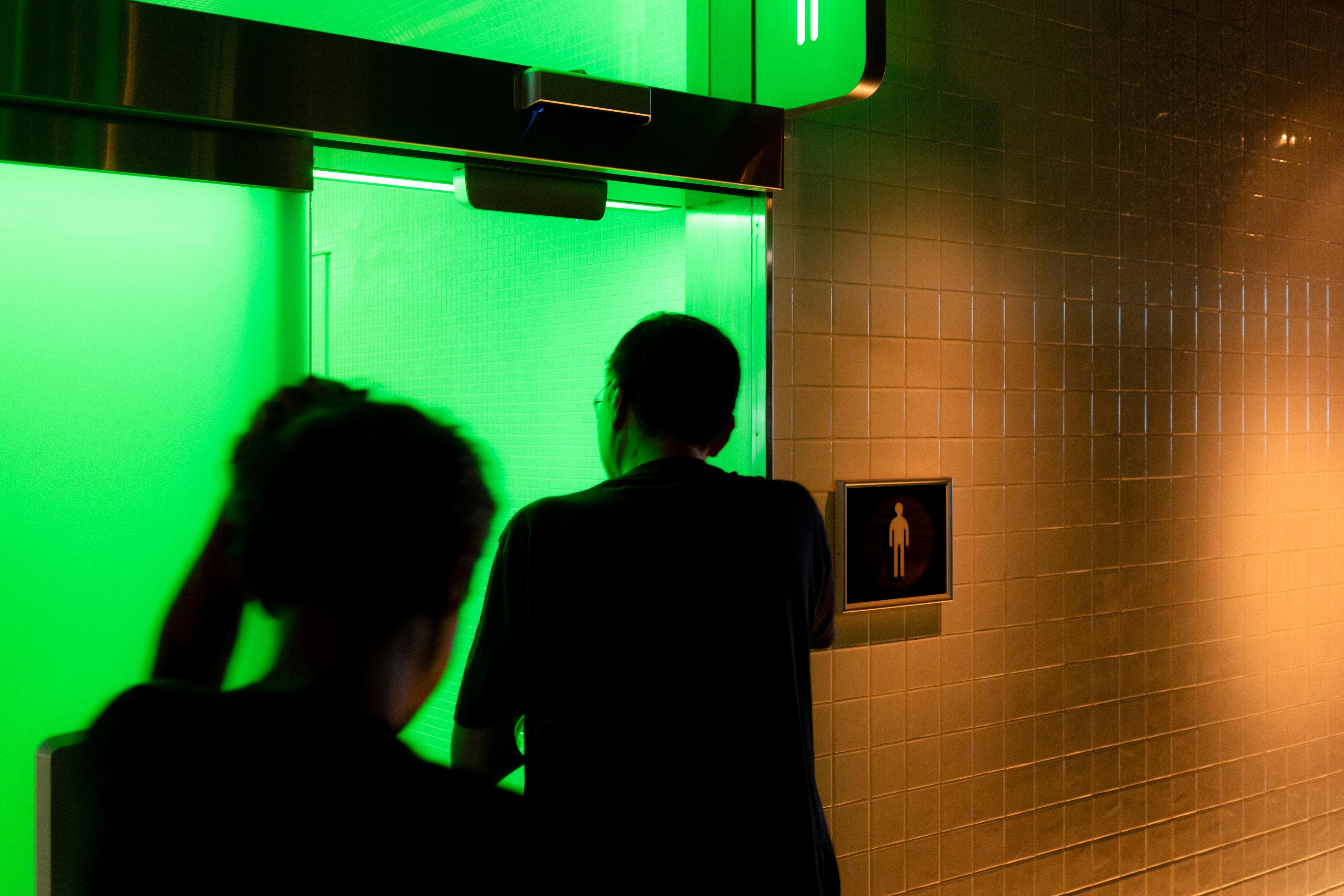 Two people near a restroom entrance illuminated by green light. A male restroom sign is mounted on the adjacent tiled wall. One person stands close to the door, partially open, while the other is closer to the viewer.