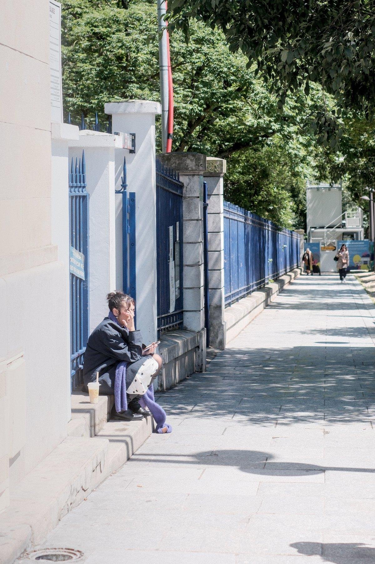 Photo verticale. Une femme brune cheveux en petit chignon négligé, en longue veste bleu, long foulard violet, assise sur le rebord d’une grille bleu (qui continue vers le point de fuite, section par section après une interruption avec un linteau montrant une entrée de parc), téléphone dans la main gauche, bubble tea posé à sa droite
