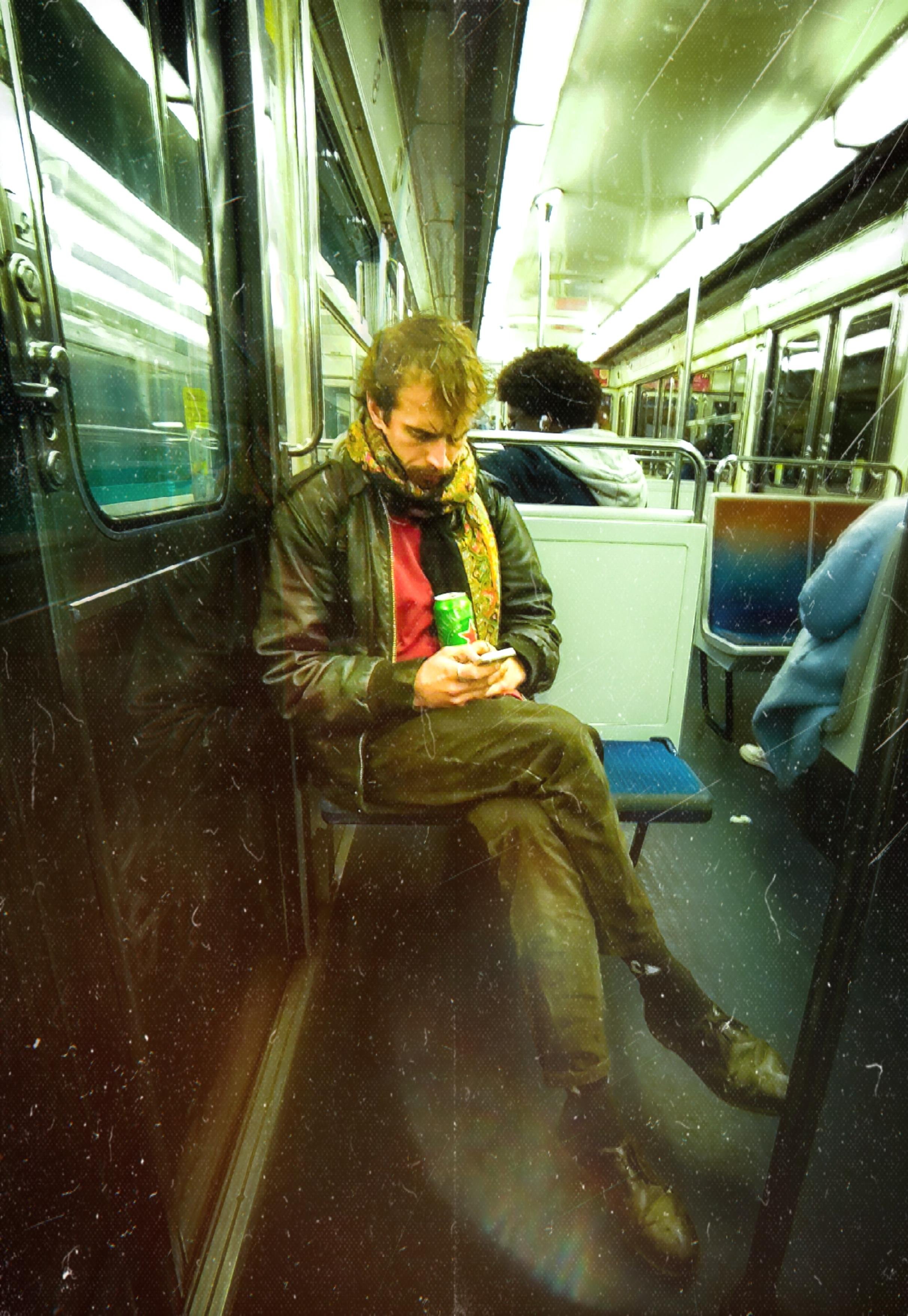 L'image montre une personne assise dans un métro, tenant une canette et regardant son téléphone. La personne porte une veste en cuir et une écharpe. L'intérieur du train est visible, avec un éclairage fluorescent et d'autres passagers assis à proximité.