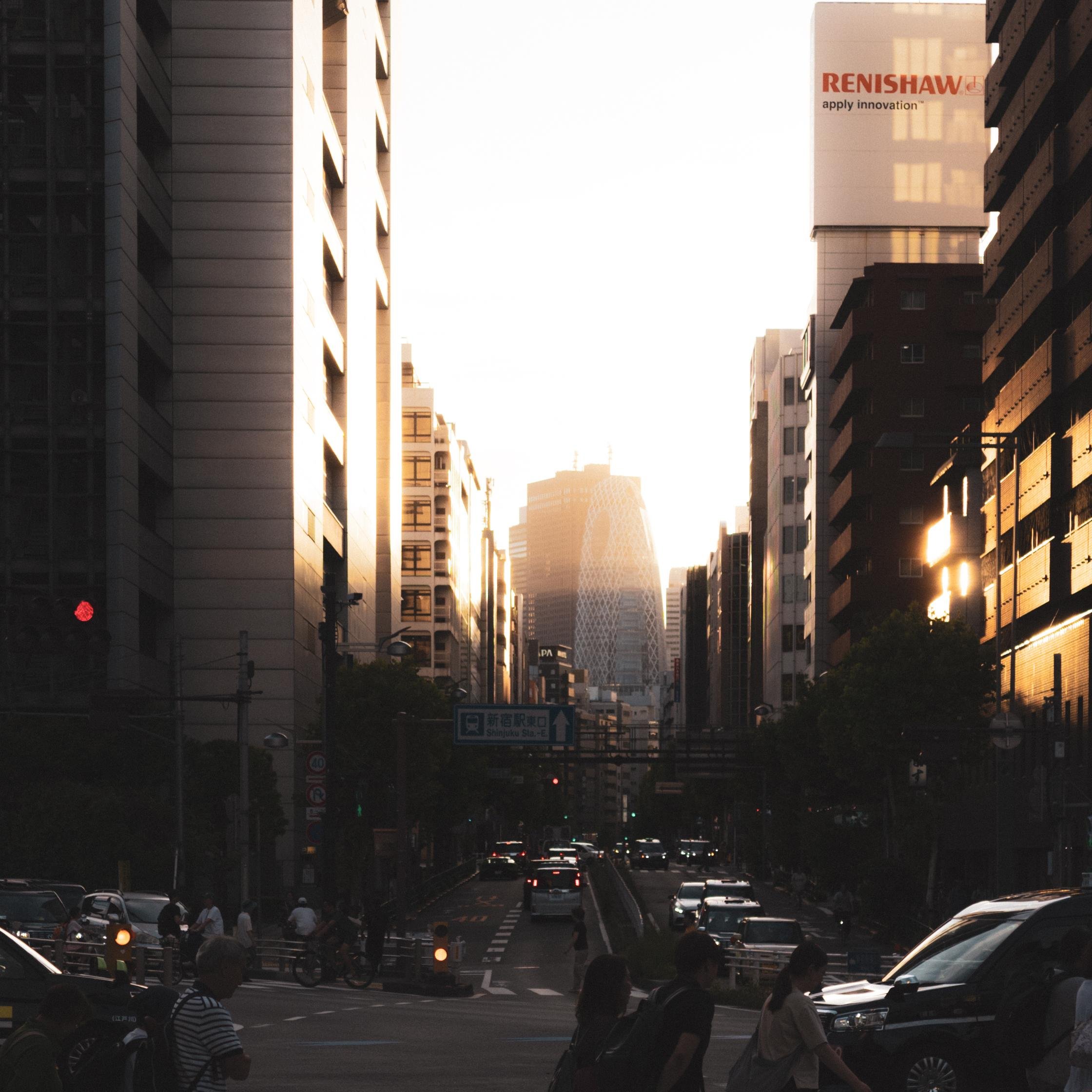 Urban street scene at sunset with tall buildings on either side. The sun casts a warm glow, highlighting windows and structures. Pedestrians and vehicles move along the street, while distant skyscrapers, including one with a distinctive mesh pattern, are visible in the background. A traffic light is red, and a sign with the word "RENSHAW" is on a building.